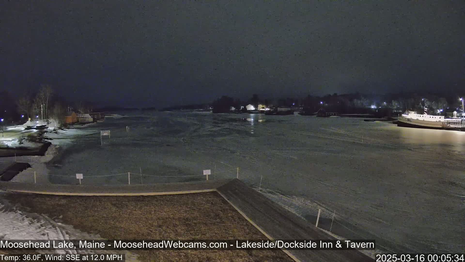 A nighttime view of a partially frozen lake with a boat docked on the far shore, under a dark, clear sky.