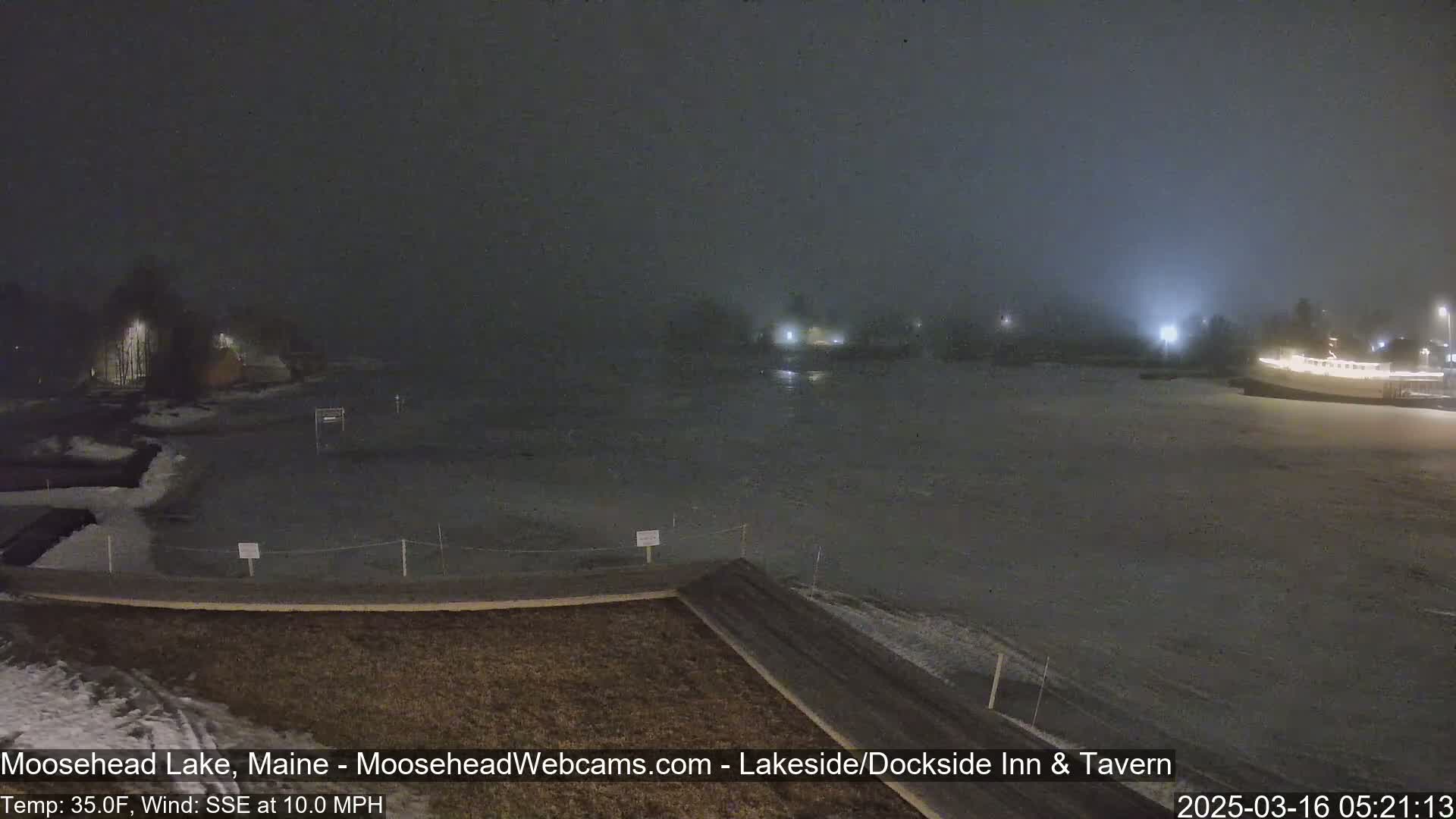 A nighttime, foggy view of a snow-covered lake and shoreline with some lights visible in the distance, under a 10 mph wind.