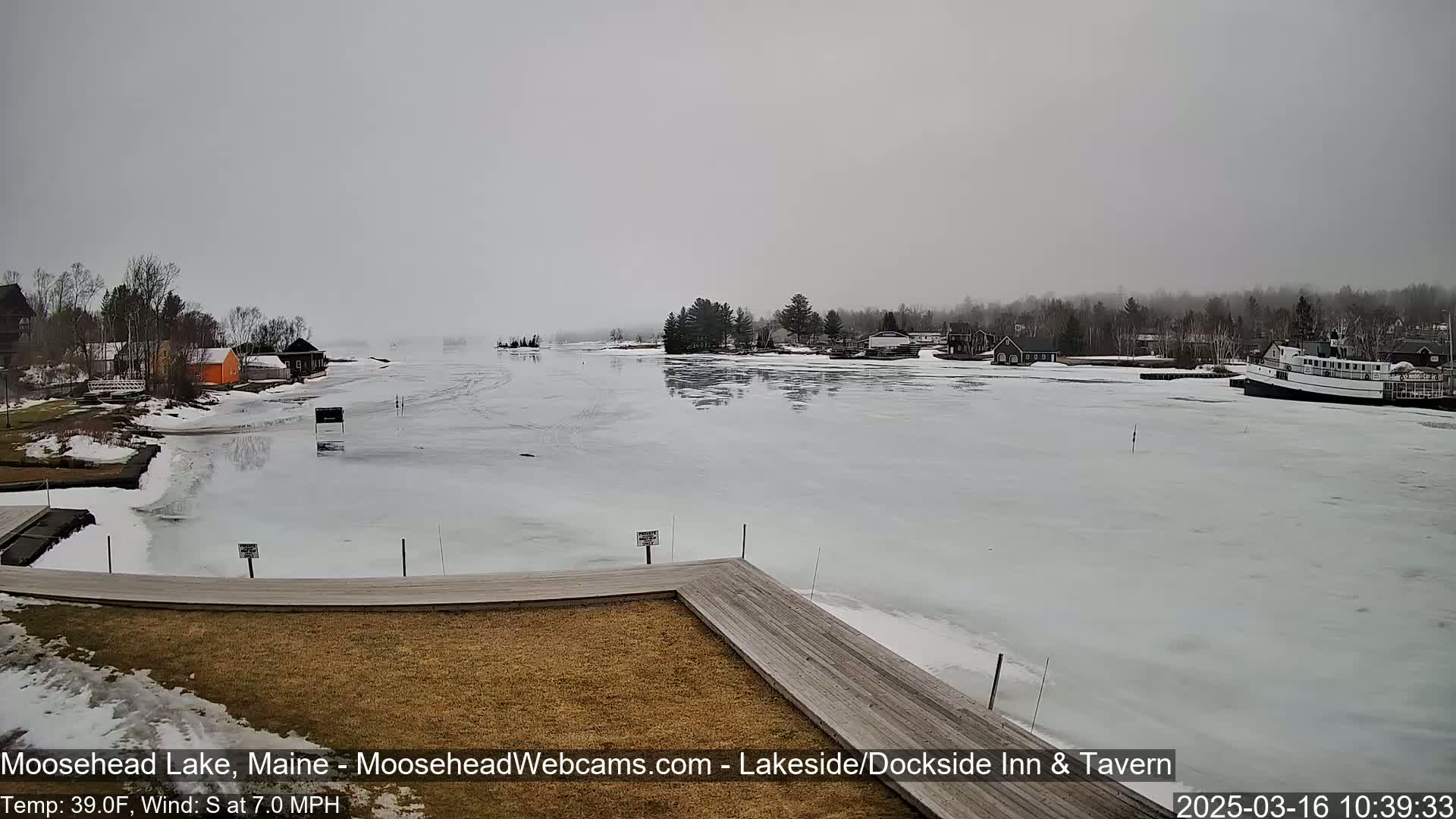 A partially frozen lake is seen from a dock on an overcast day, with a few buildings and a boat visible in the distance.