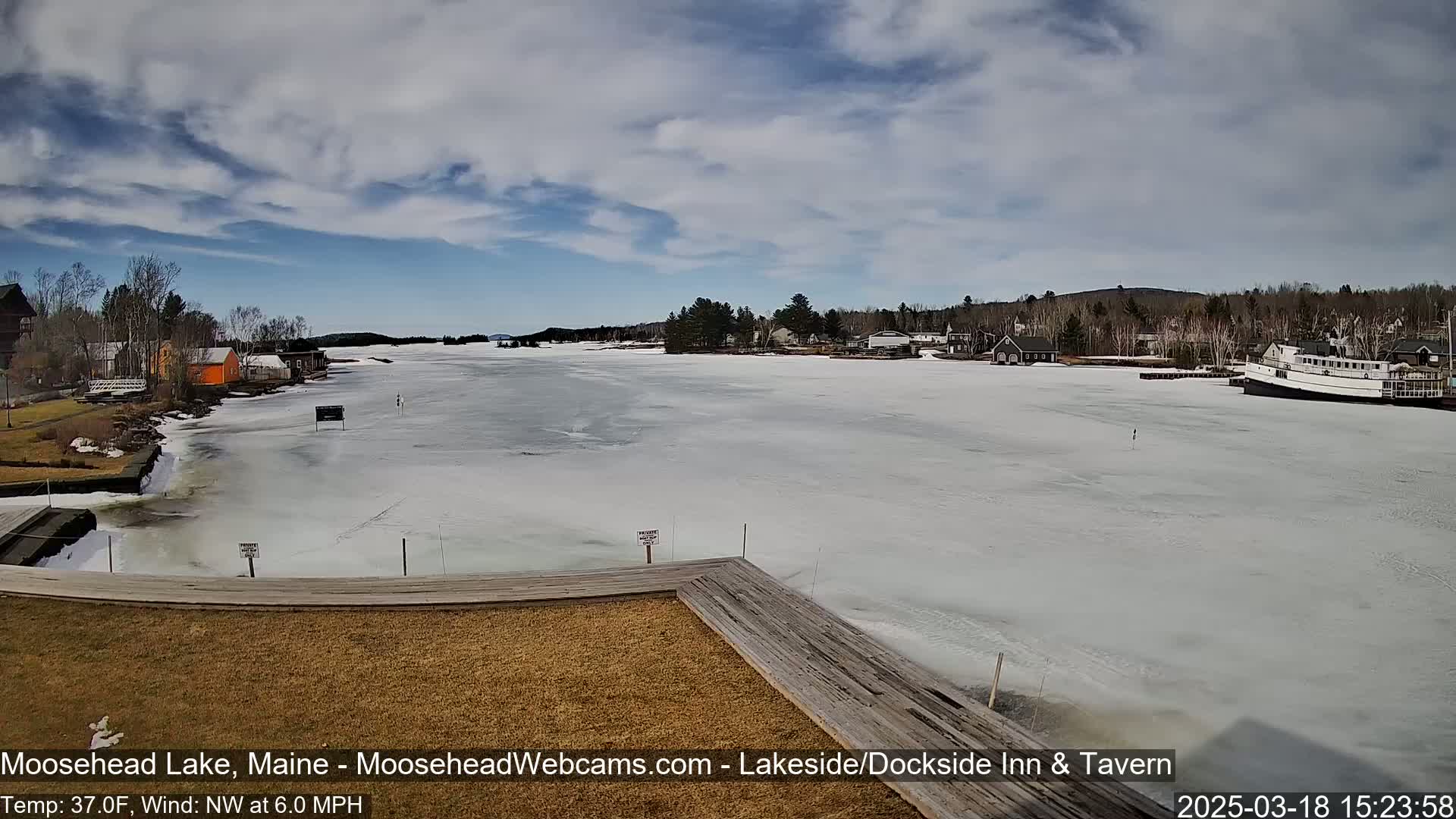 A partially frozen lake is viewed from a wooden dock, with buildings and trees lining the shore under a partly cloudy sky.