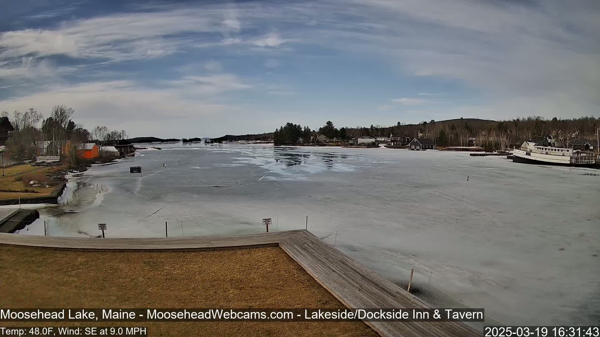 A partially frozen lake with buildings and a boat on the shore under a partly cloudy sky.