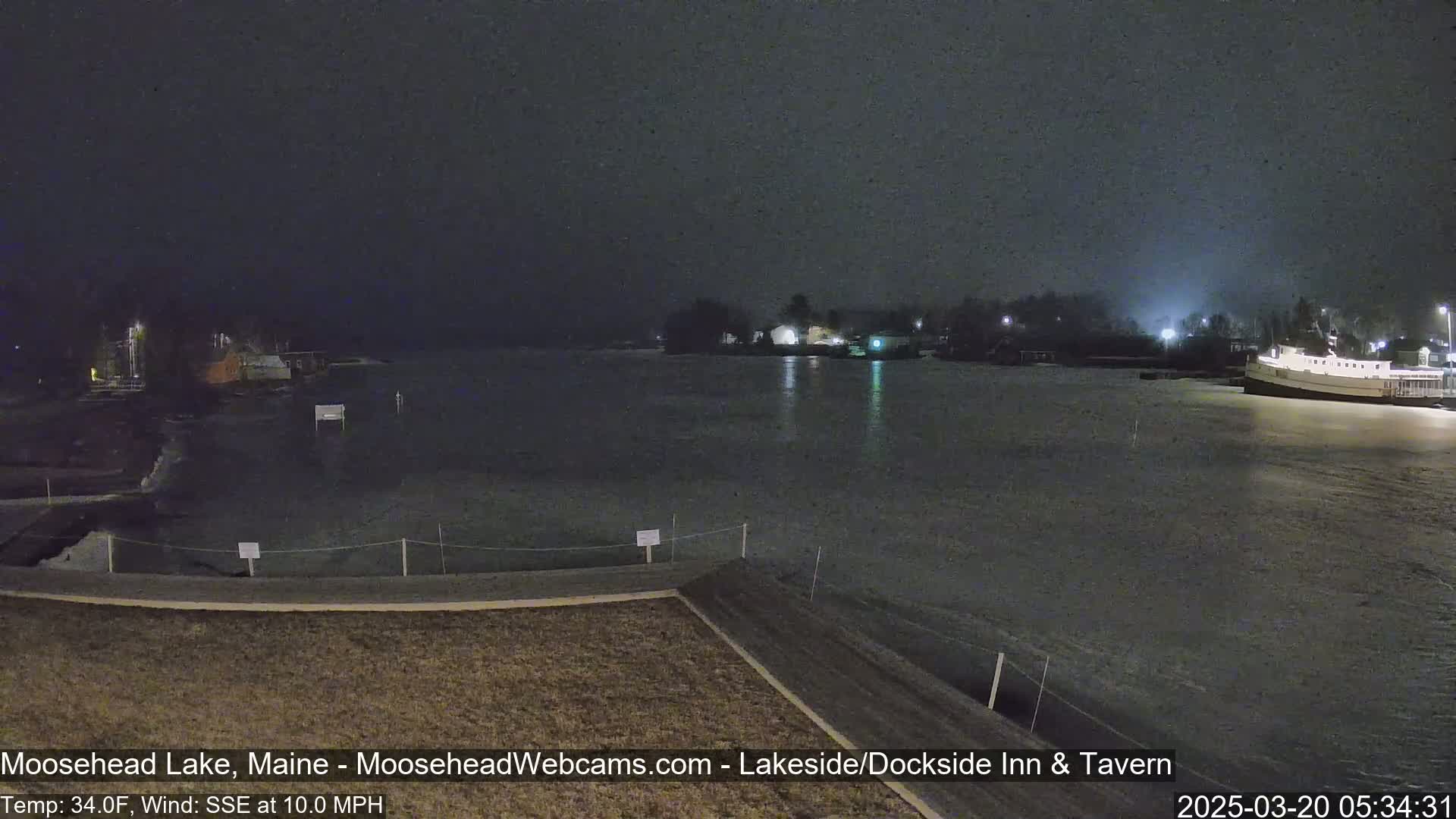 A nighttime view of a partly frozen lake with a few lights visible on the shore under a dark, possibly snowy sky.