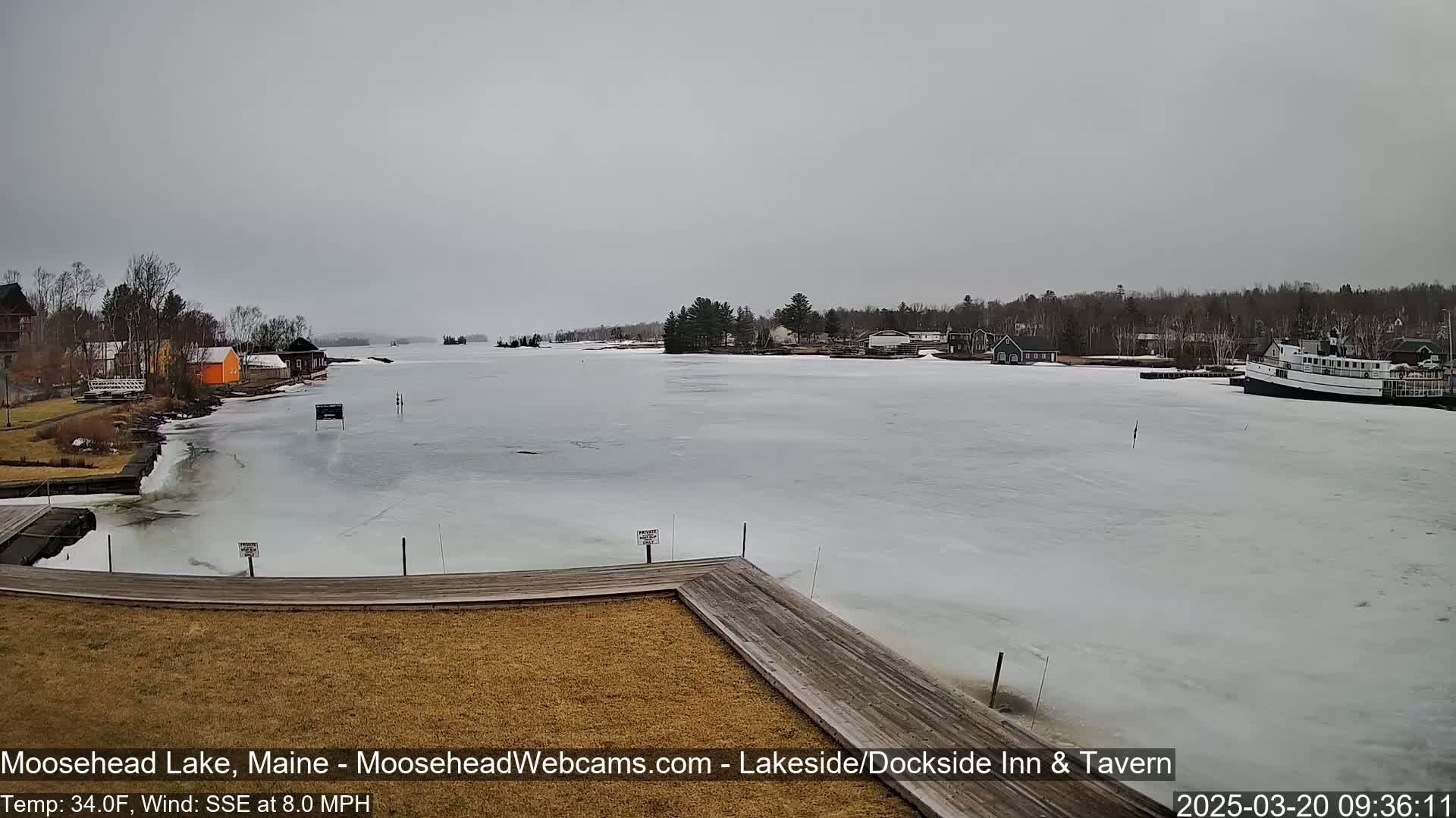 A mostly frozen lake is seen from a dock, with a few buildings and a boat visible on the shore under an overcast sky.