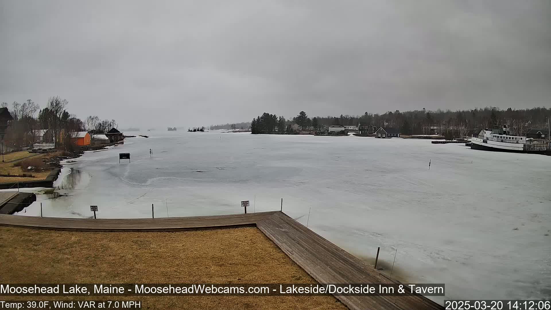 A partially frozen lake with buildings and trees along the shoreline under an overcast sky.