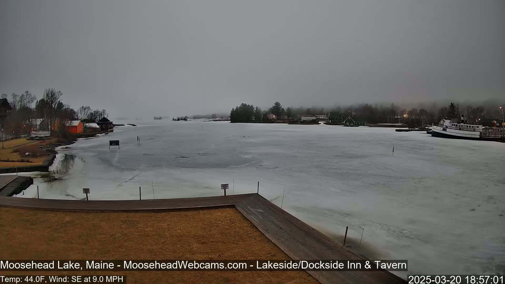 A partially frozen lake is seen from a dock on an overcast day, with buildings and a boat visible in the distance.