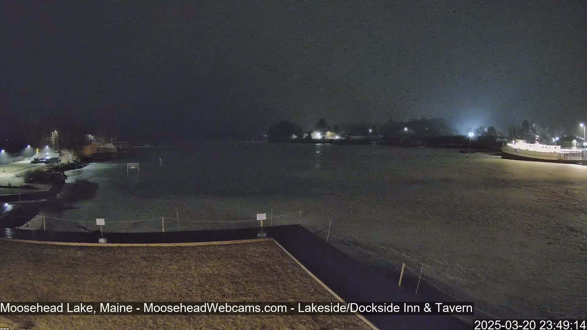 A nighttime view of a snow-covered lakeshore with buildings in the distance under a lightly snowing sky.