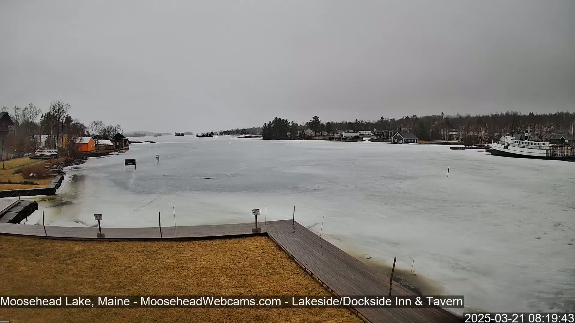 A partially frozen lake is seen from a dock on an overcast day, with buildings and trees visible along the shoreline in the distance.