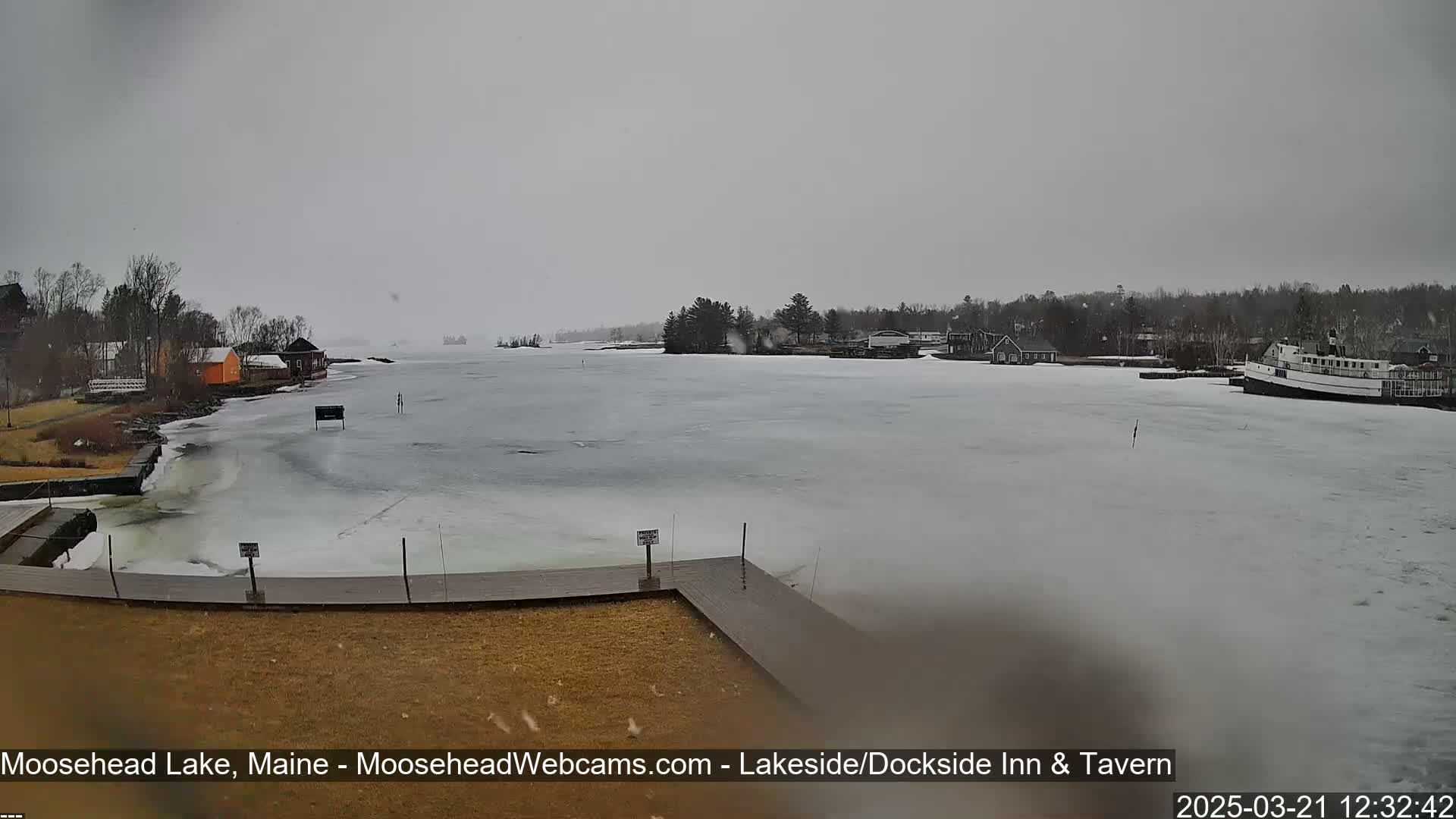 A partly frozen lake is seen on an overcast day, with a docked boat and shoreline buildings visible in the distance.