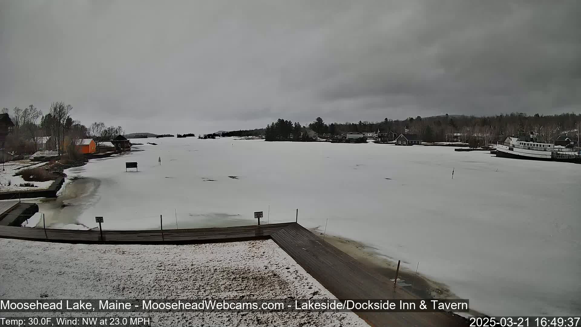 A mostly frozen lake, under an overcast sky, is seen from a snow-covered dock with a few buildings and a boat visible in the distance.