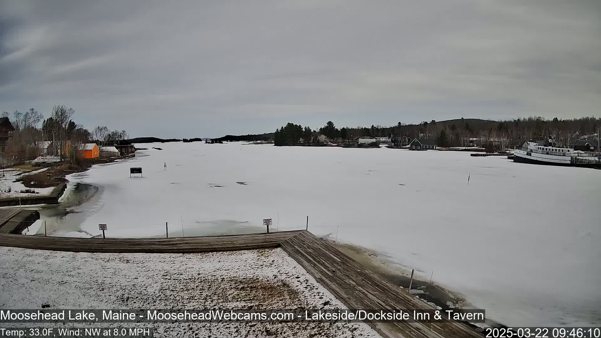 A mostly frozen lake with a few buildings and a boat visible in the distance under an overcast sky.