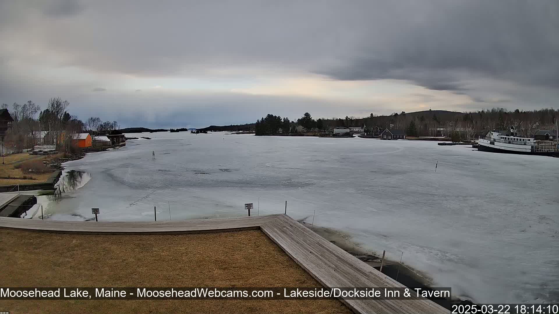 A mostly frozen lake is seen from a lakeside dock on an overcast day, with buildings and trees lining the shore.