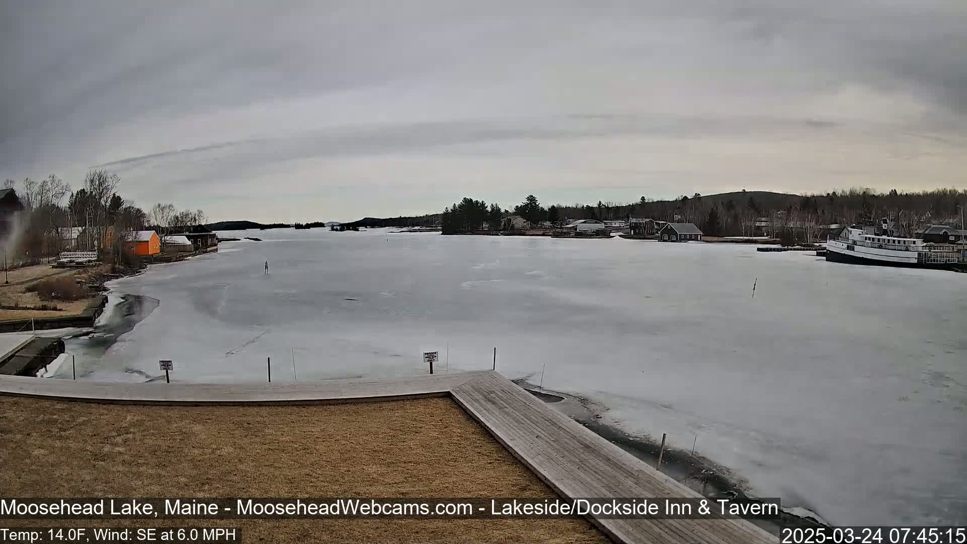 A partially frozen lake is seen from a wooden dock on an overcast day, with buildings and trees visible along the shoreline.