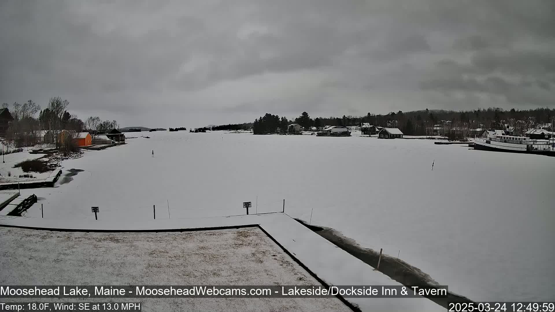 A snow-covered lake stretches out to a treeline under an overcast sky, with a few buildings and a boat visible in the distance.