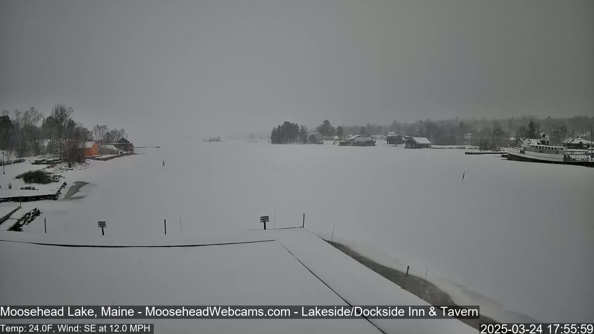 A snow-covered frozen lake is seen with buildings and trees in the background during a snowstorm with a southeast wind.
