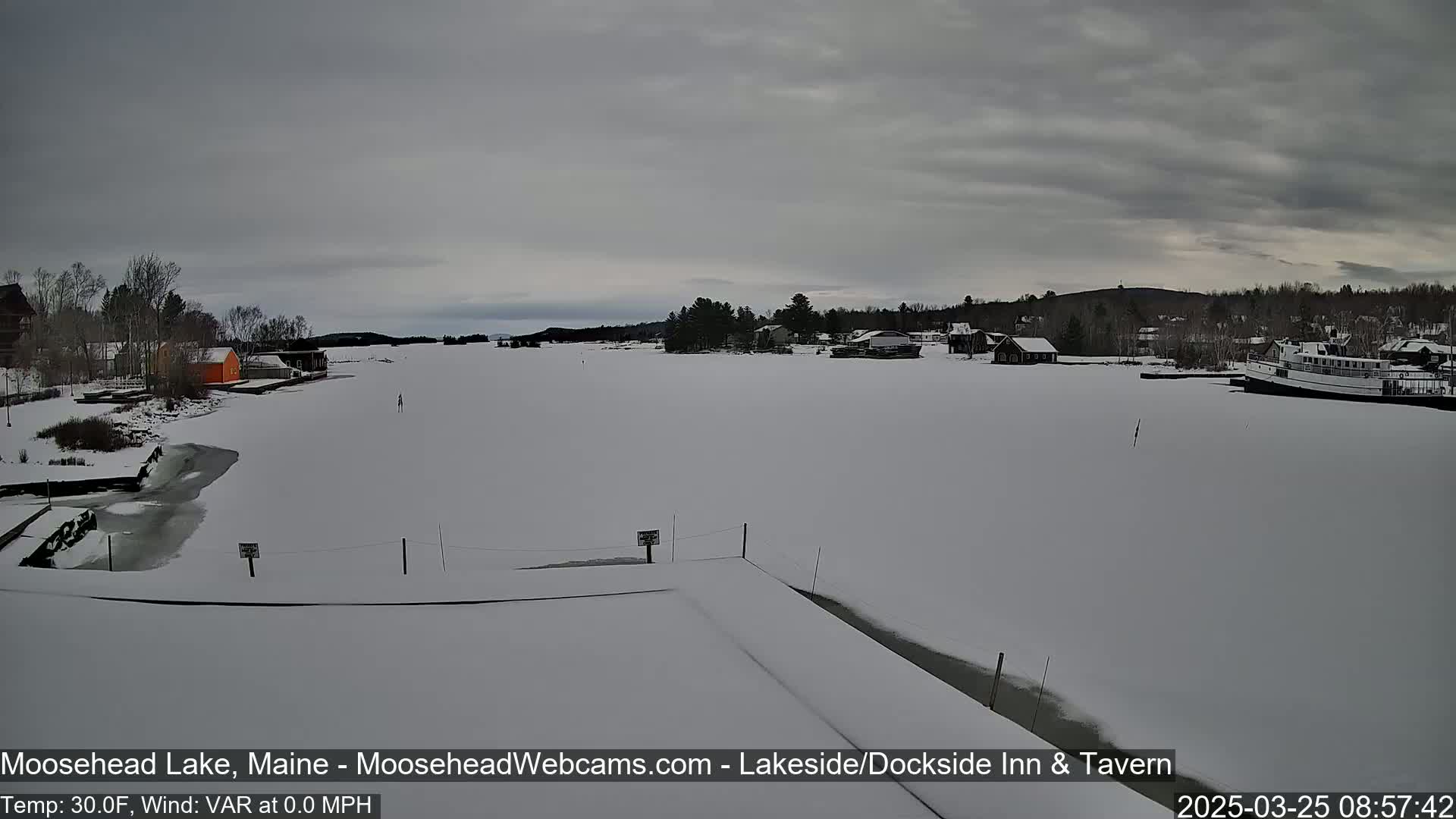 A snow-covered lake with a few buildings and a boat visible on the shore under a cloudy sky.
