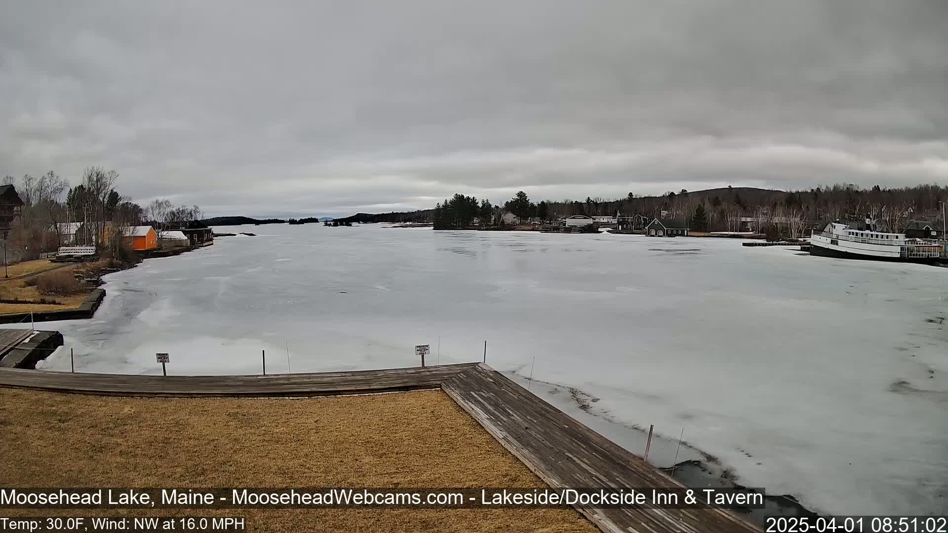 A partly frozen lake is viewed from a wooden dock under an overcast sky, with buildings and a boat visible in the distance.