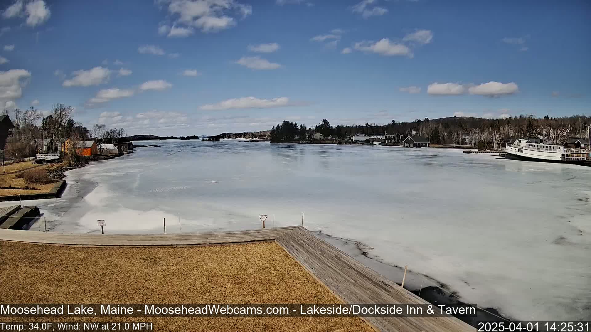 A partly sunny day shows a partially ice-covered lake with buildings and a boat on the shore, a wooden dock extending into the water, and a light northwest wind.