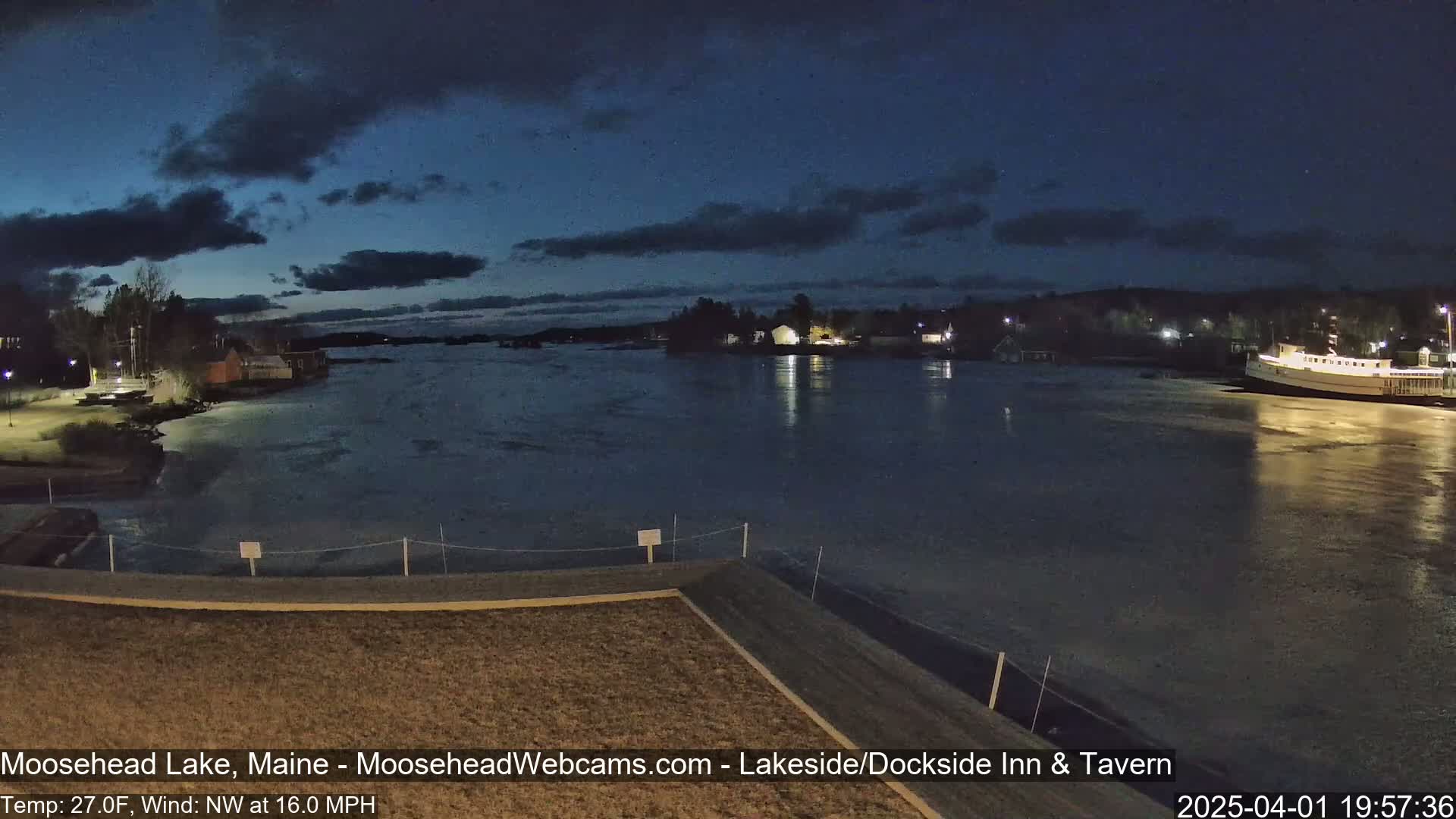 A dark, nighttime view of a calm lake partially covered in ice, with buildings and a boat visible along the shoreline under a mostly cloudy sky.