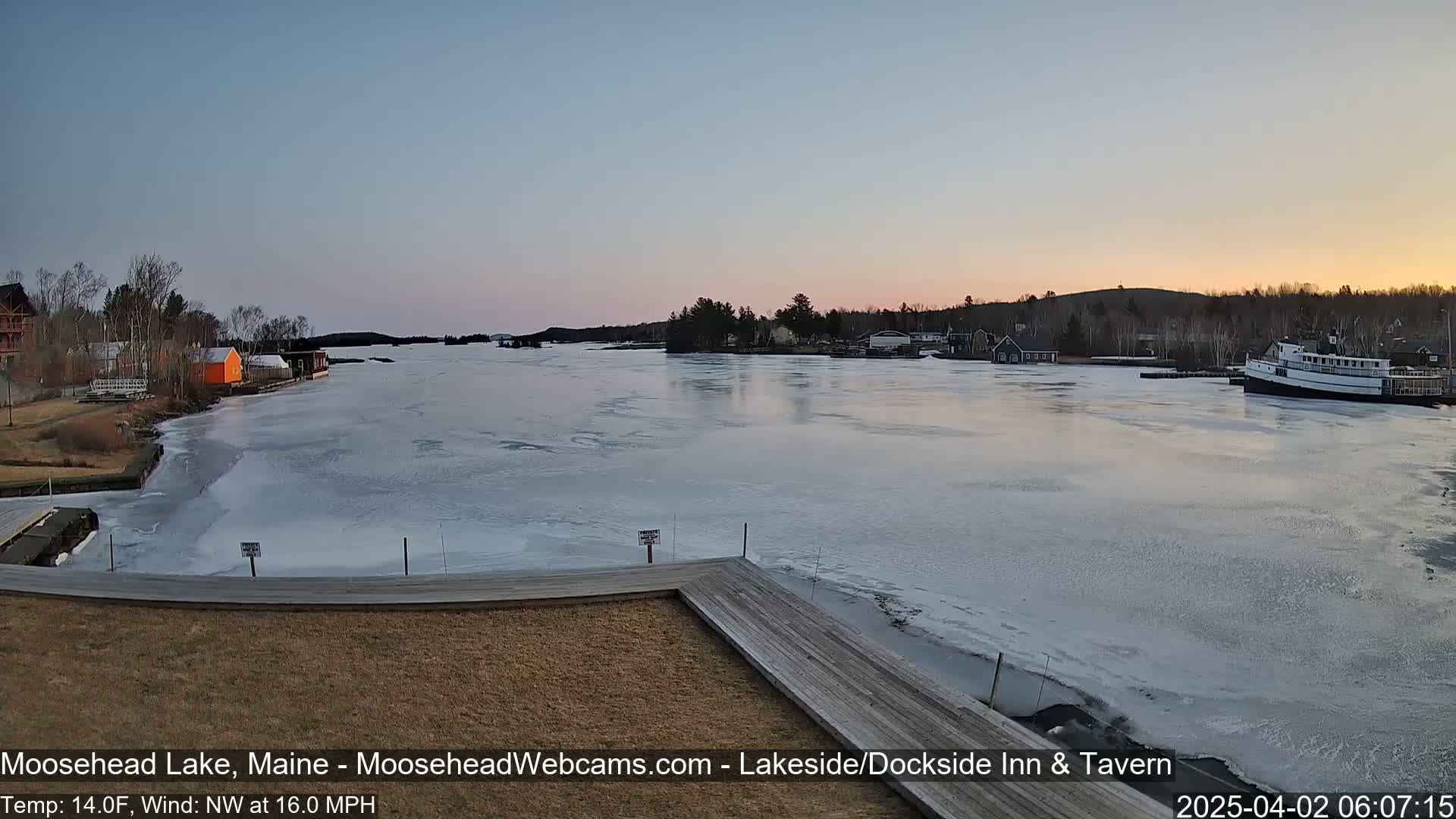 A partially frozen lake at sunset, with buildings and a boat visible on the shore, under a calm sky.