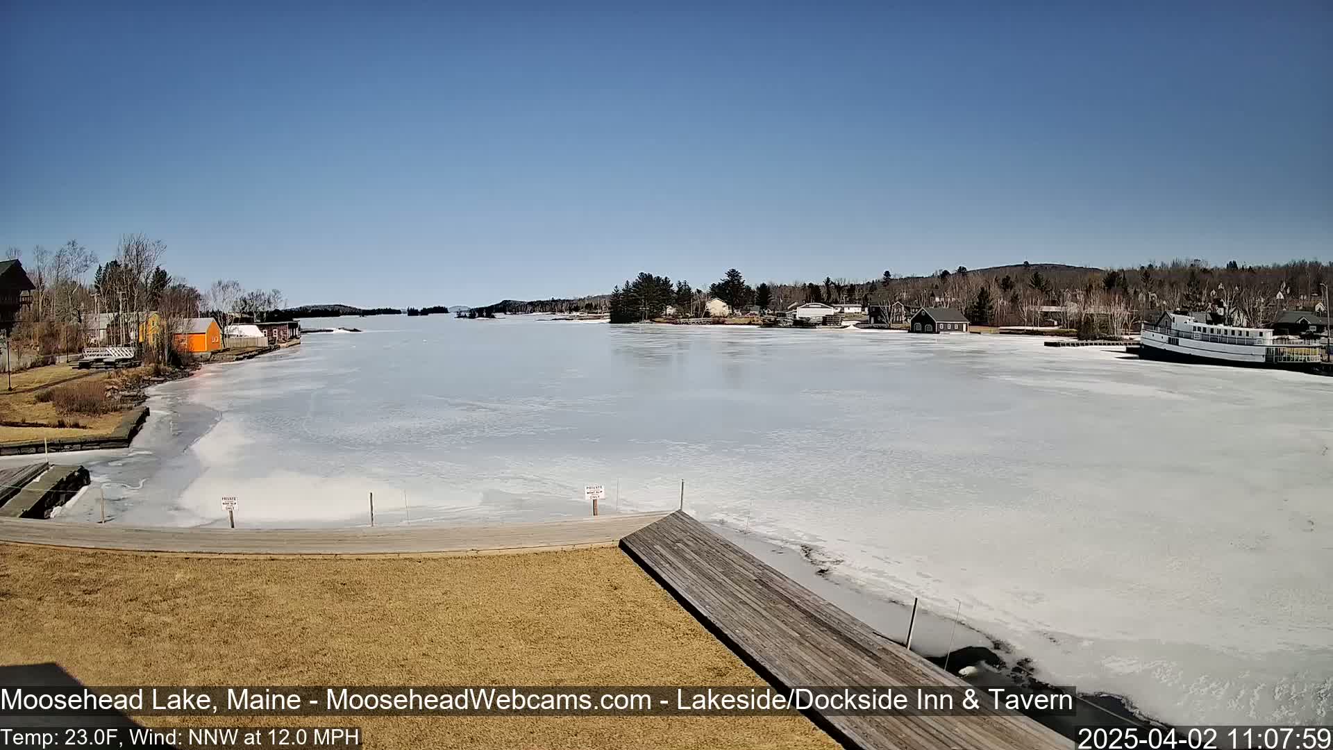 A partially frozen lake with buildings and a boat on the shore under a clear blue sky.