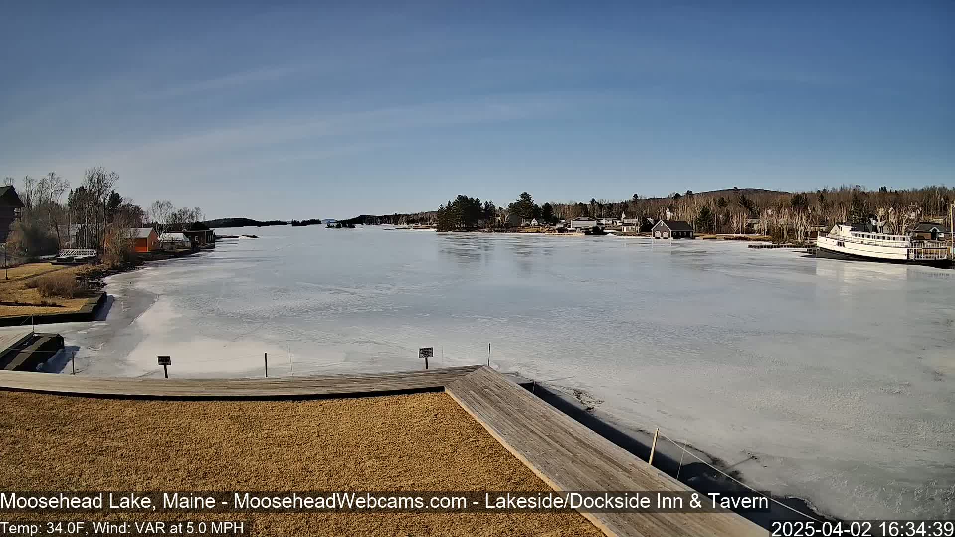 A partially frozen lake is seen from a wooden deck, with buildings and a boat visible in the background under a clear blue sky.