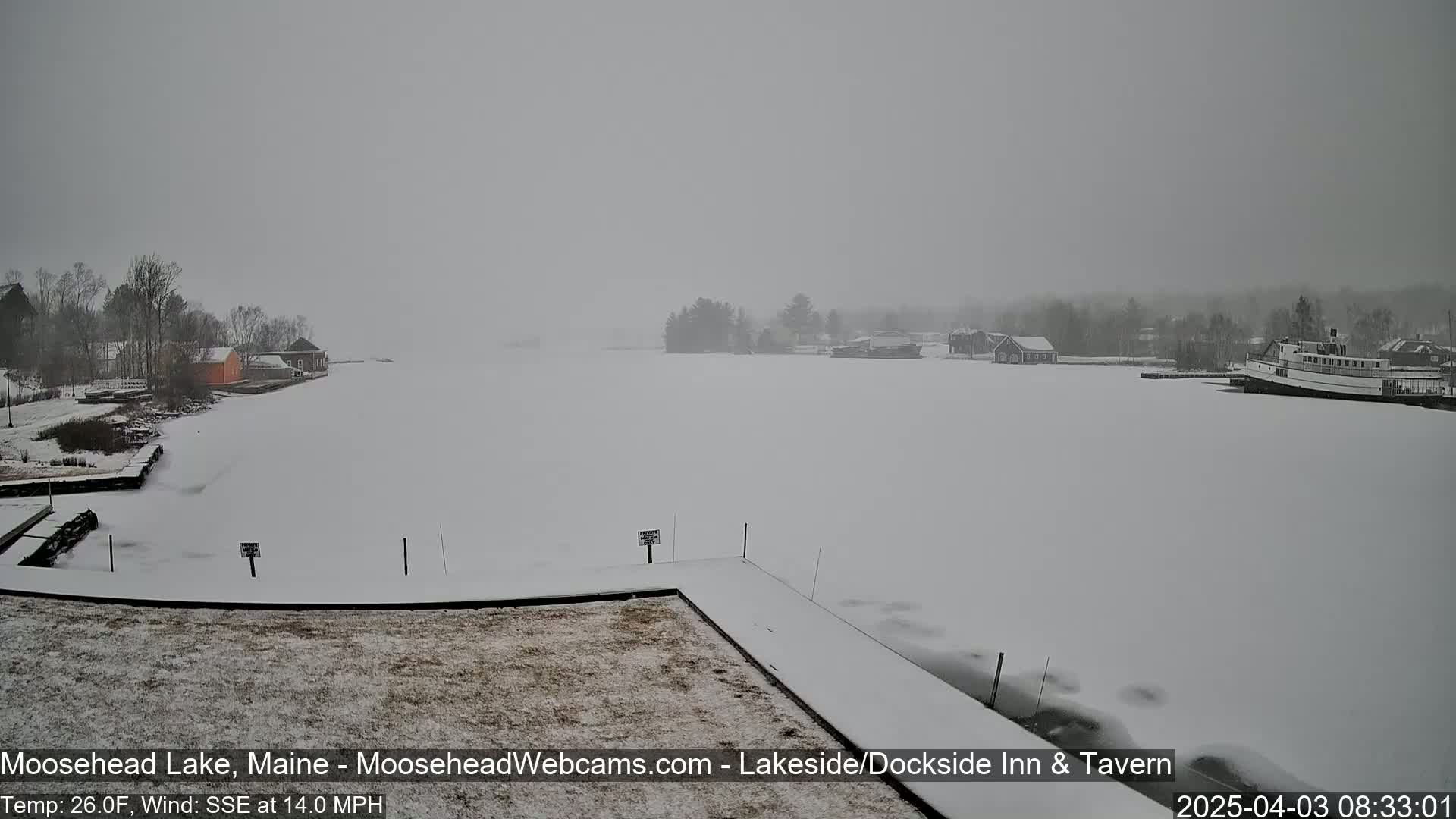 A snow-covered lake with several buildings and a boat on the far shore in snowy conditions.