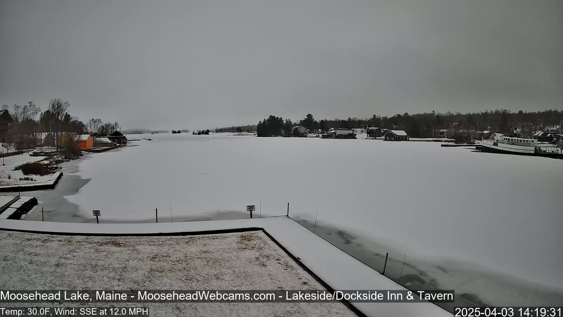 A snow-covered lake with a few buildings and a boat on the far shore is visible under an overcast sky.