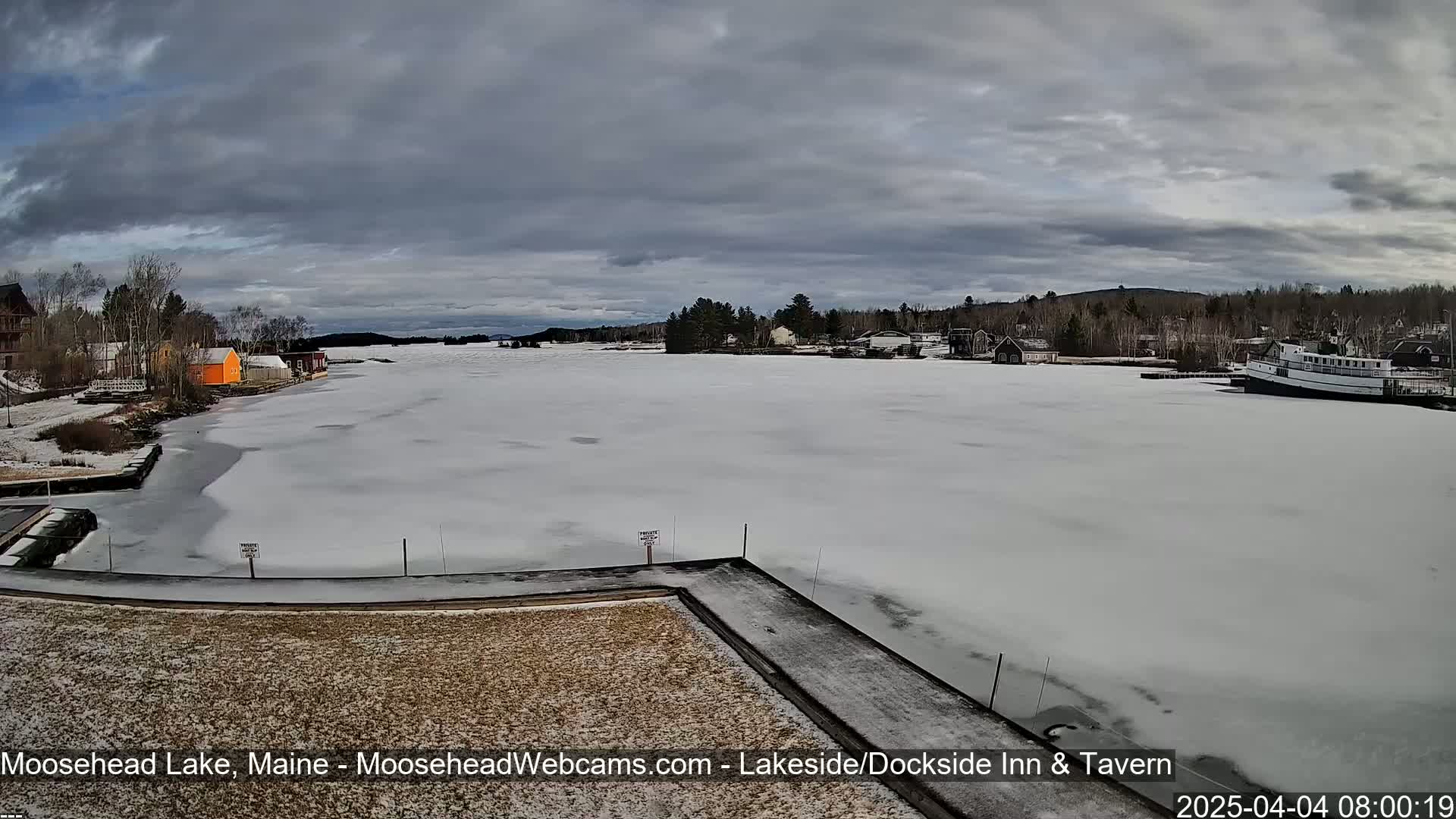 A mostly frozen lake with buildings and trees along the shore under a cloudy sky.