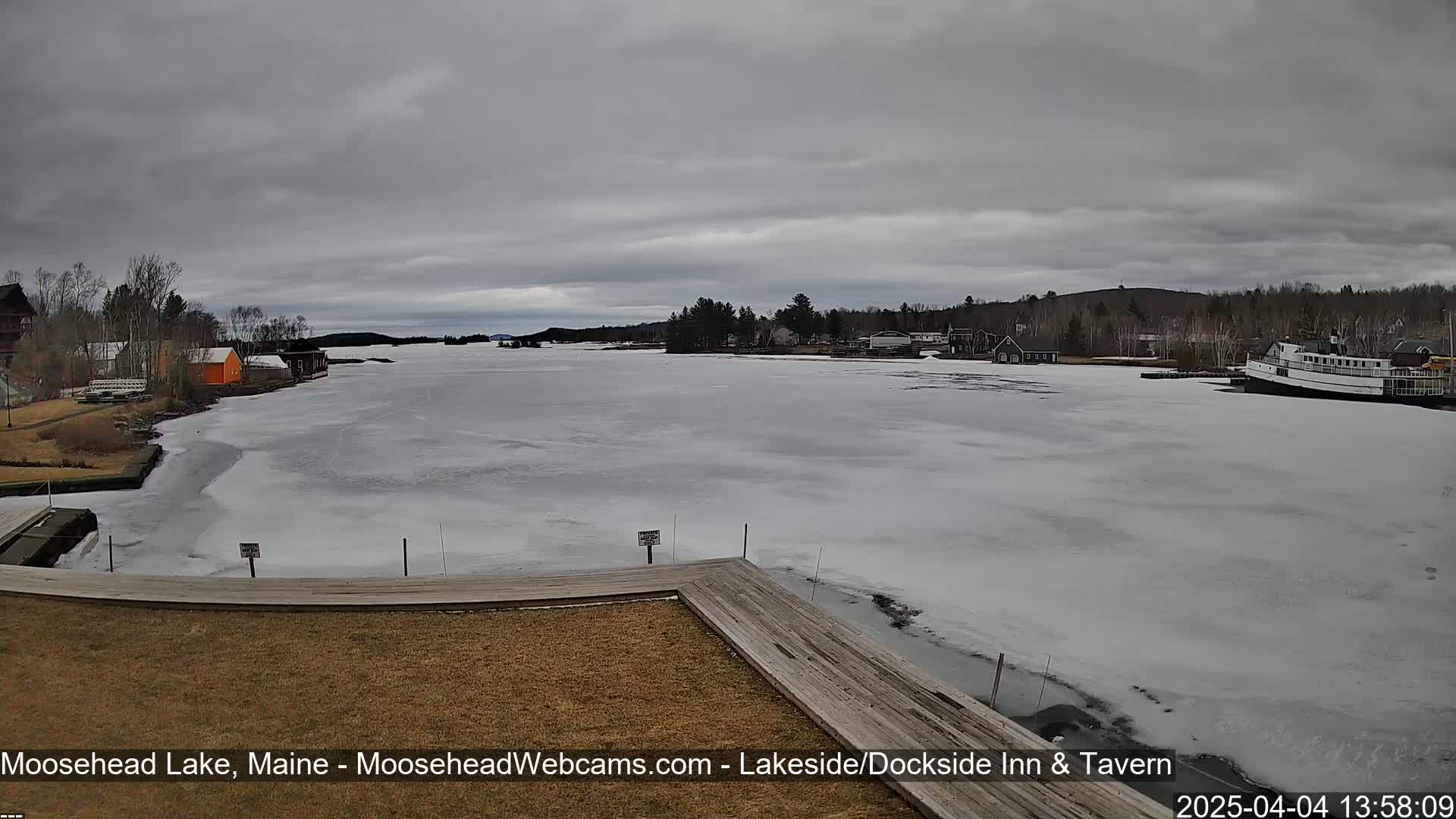 A partially frozen lake is viewed from a wooden dock on an overcast day, with buildings and a boat visible in the distance.