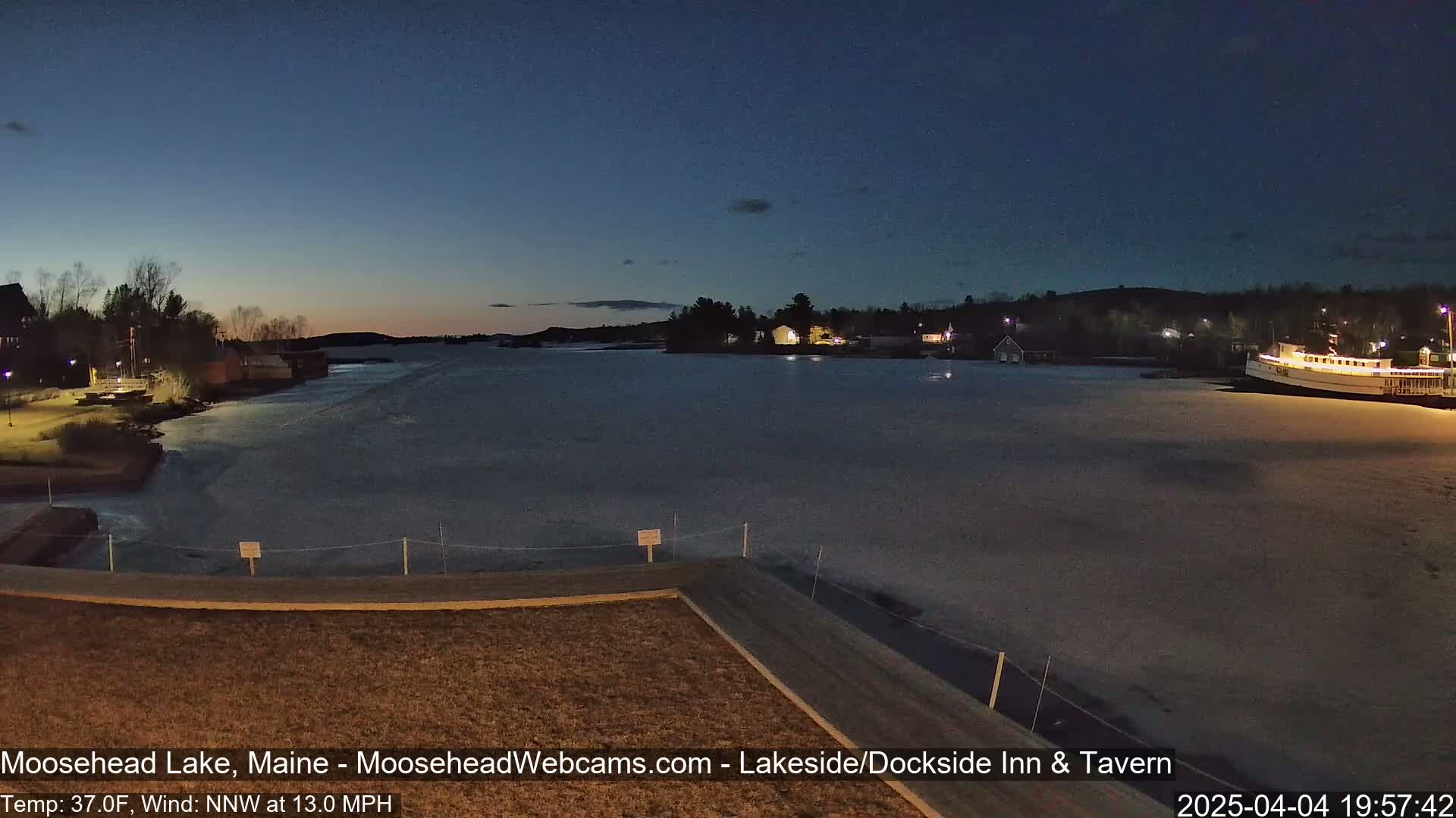 A partially frozen lake at twilight, with buildings and a boat visible along the shore under a clear, dark sky.