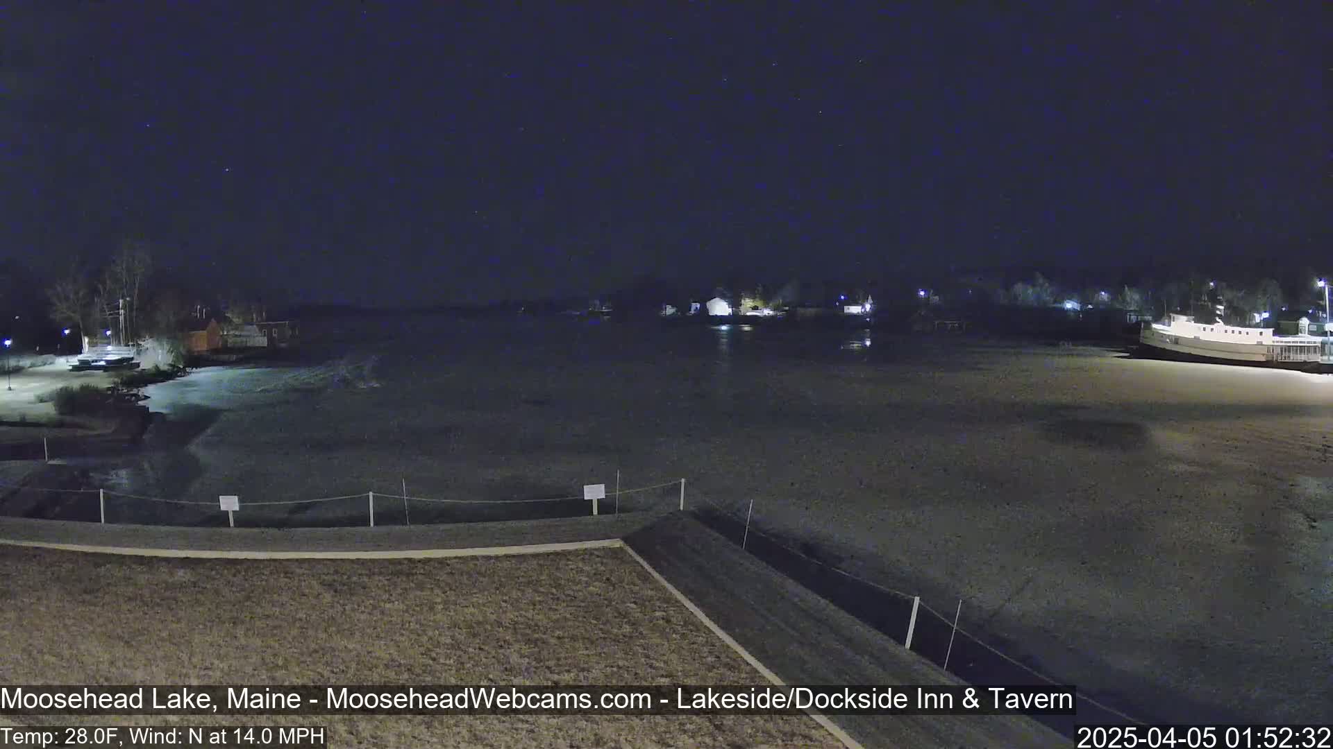A nighttime view of a partially frozen lake with buildings and a boat visible in the distance under a clear, starry sky, with a north wind blowing at 14 mph and a temperature of 28°F.