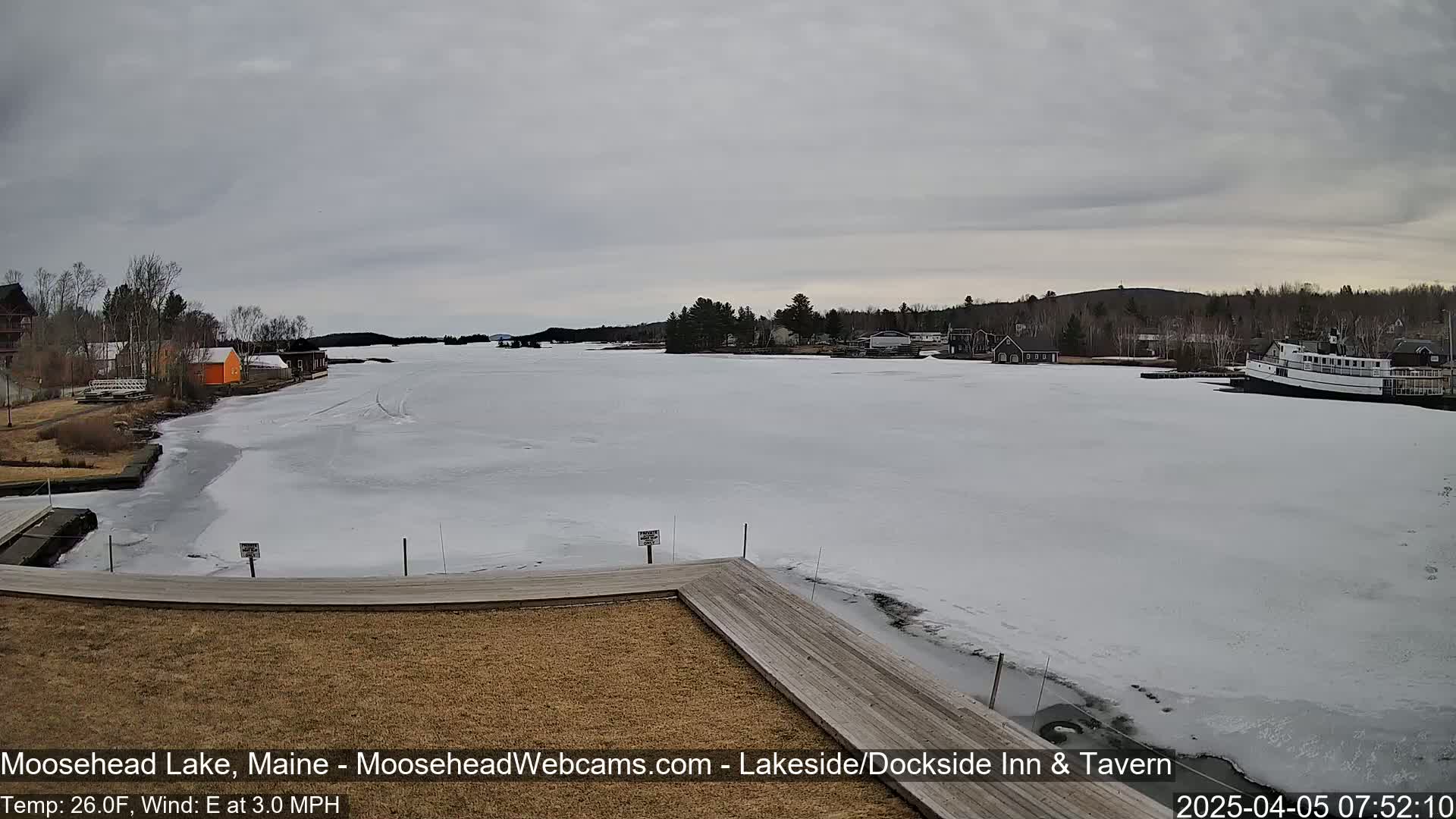 A partially frozen lake is seen from a wooden dock, with buildings and a boat visible in the distance under an overcast sky.