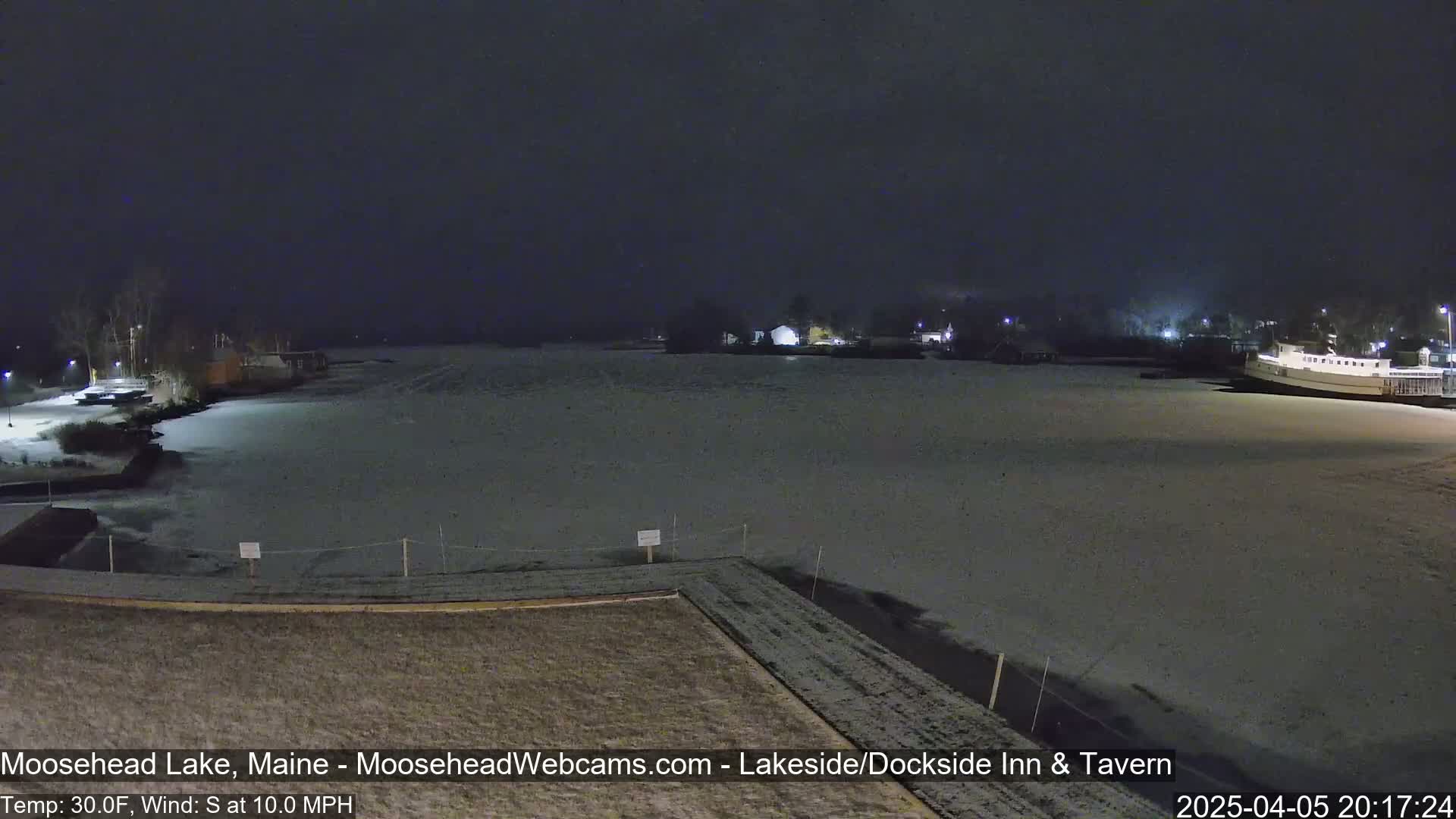 A snow-covered frozen lake is seen at night, with a few buildings and some lights visible in the distance under a dark, cloudy sky.