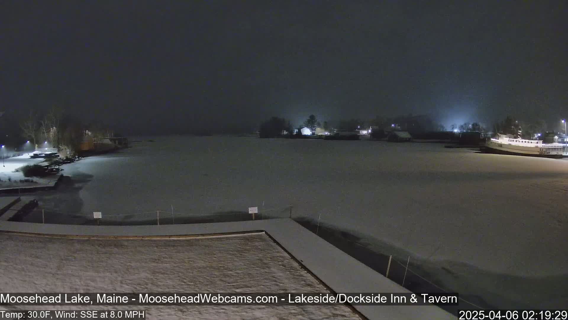 A snow-covered lake at night, with a few buildings and trees faintly visible in the distance under a dark, cloudy sky.