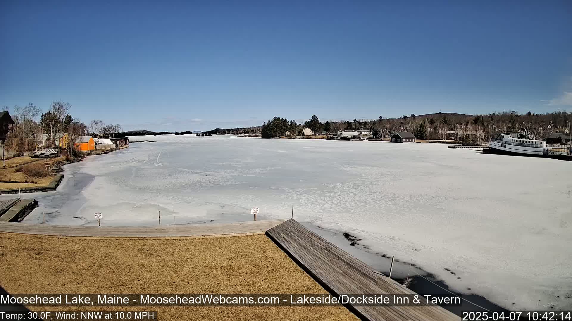 A partially frozen lake with buildings and a boat visible in the distance under a clear blue sky.