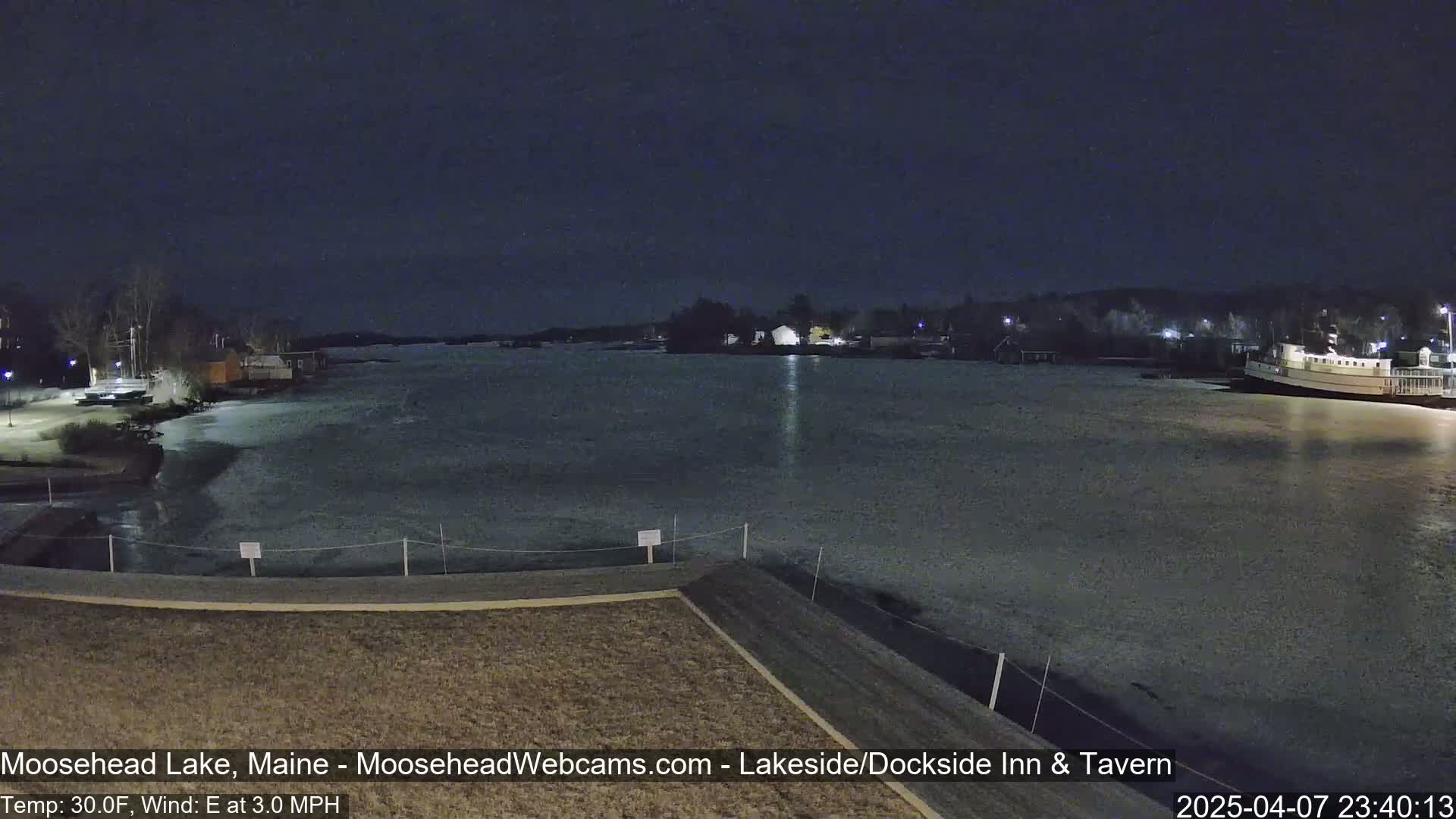 A nighttime view of a partially frozen lake with a boat docked at the far shore, under a clear, dark sky, and a light easterly wind.