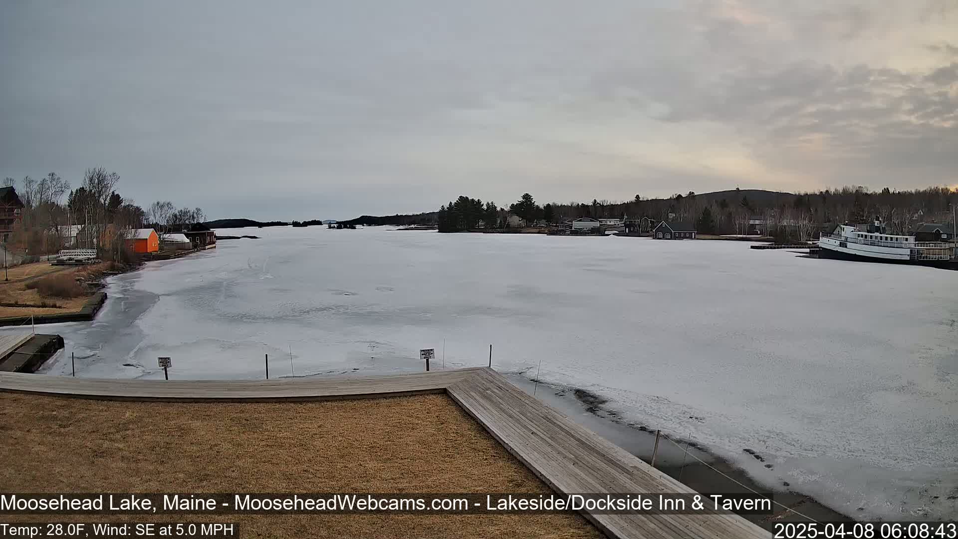 A mostly frozen lake is seen from a dock, with a few buildings and a boat visible along the shore under an overcast sky.