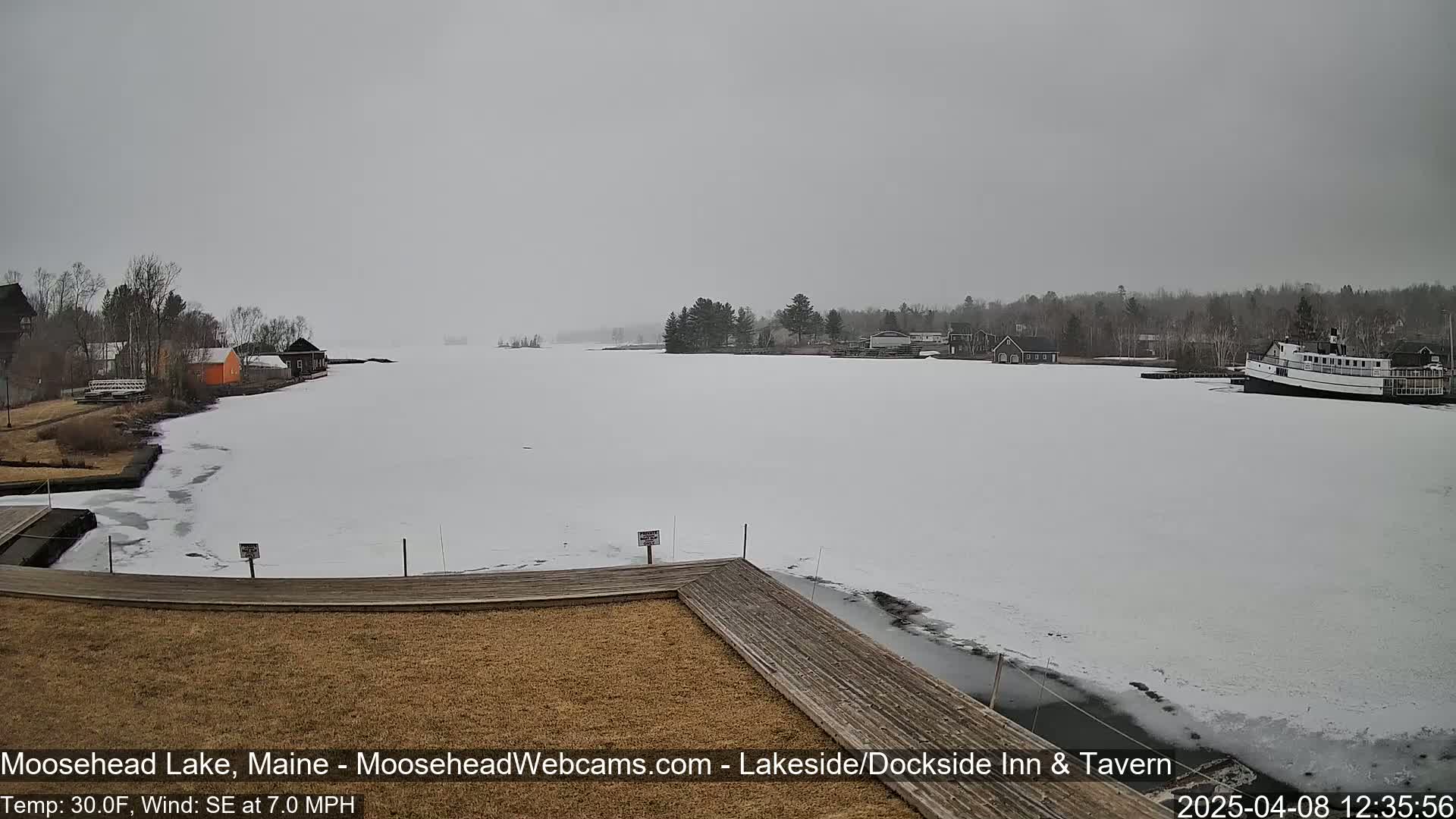 A partly frozen lake with a few buildings and a boat on the shore is visible under an overcast sky.
