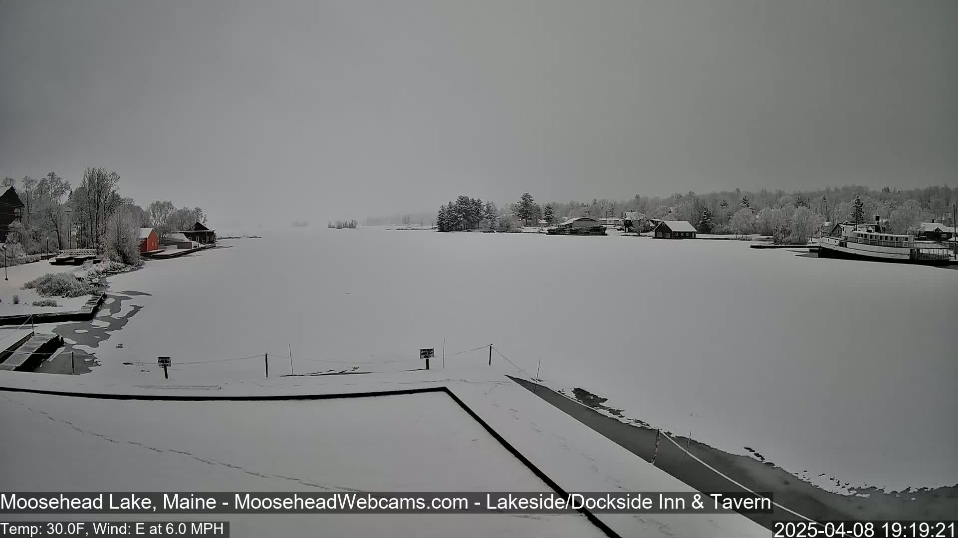 A snow-covered lake with buildings and a boat on the shore in overcast conditions.