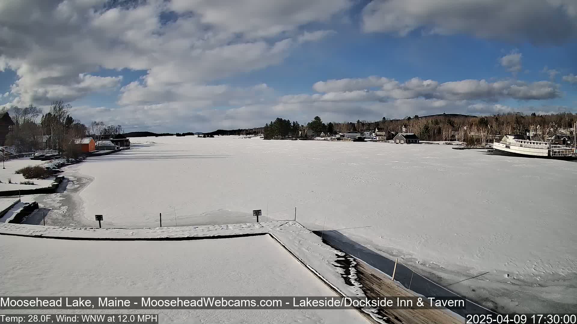 A mostly sunny day shows a frozen lake with snow-covered buildings and a docked boat in the distance.
