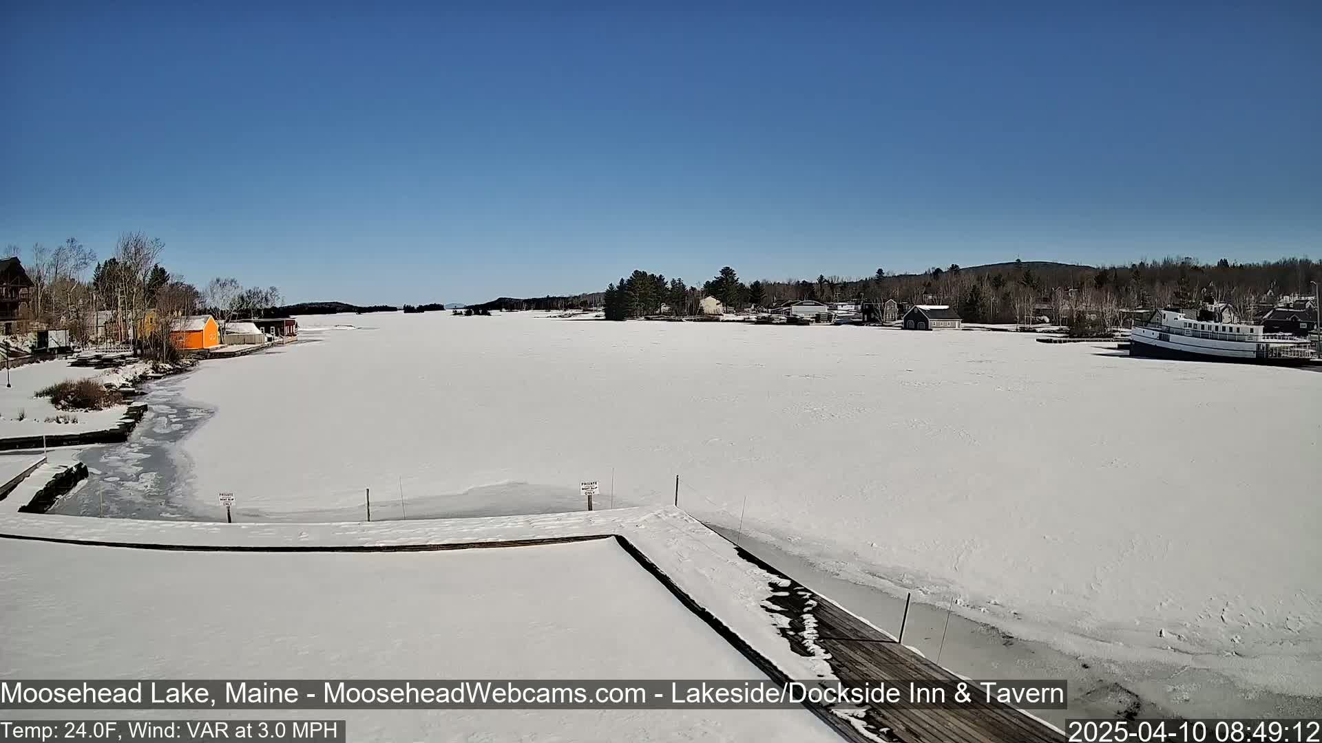 A snow-covered lake with a few buildings and a boat visible along the shoreline under a clear blue sky.