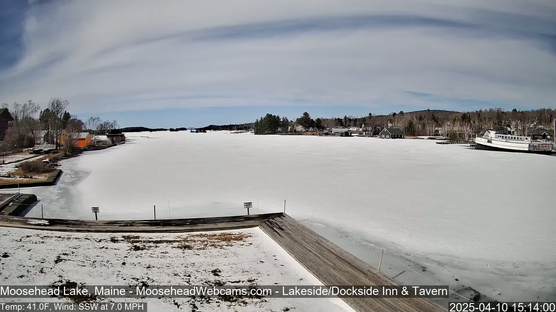 A mostly frozen lake with a few buildings and a boat visible in the distance under a partly cloudy sky.