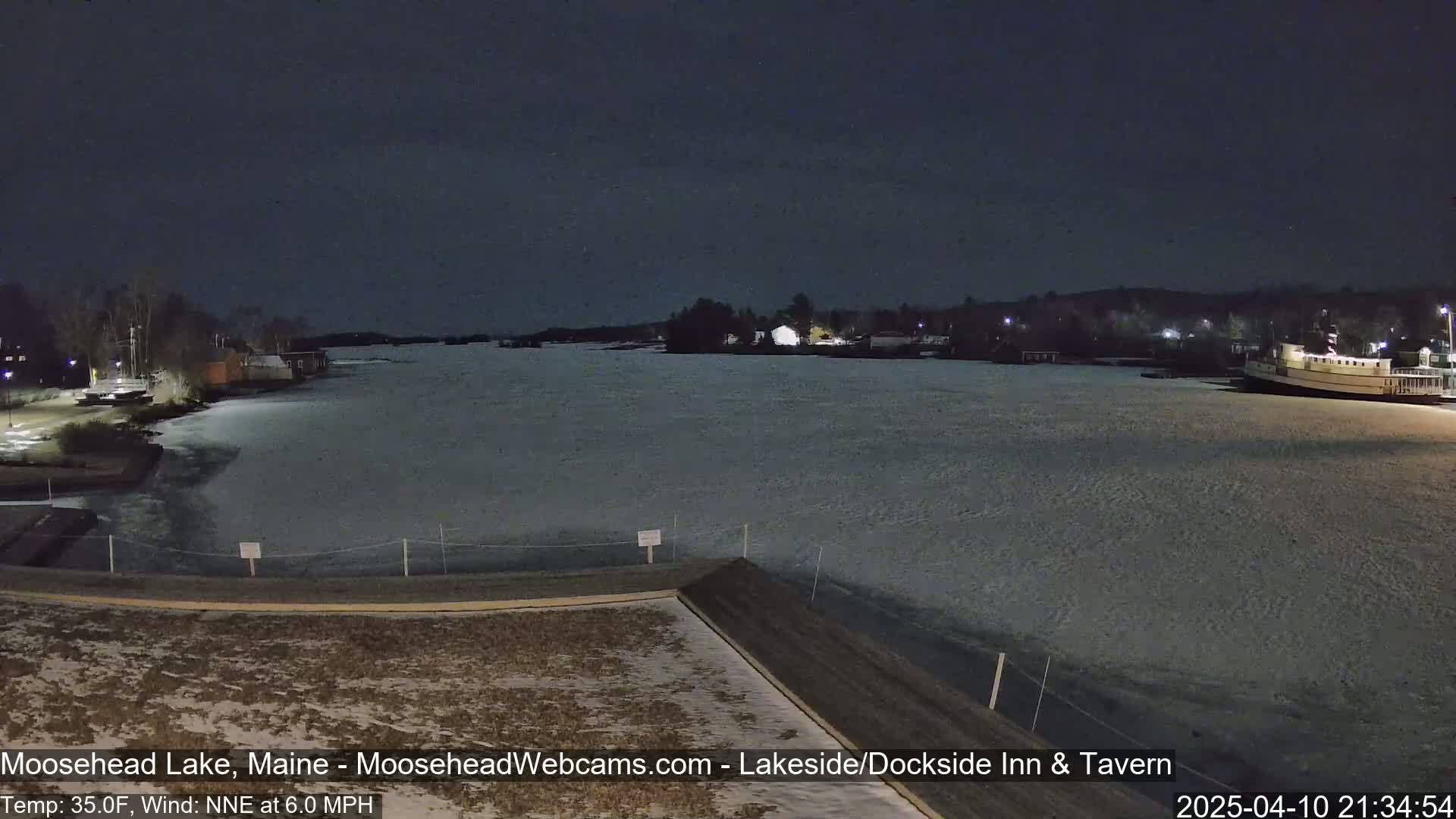 A partially frozen lake at night is viewed from a rooftop, with a boat docked on the far shore and buildings visible along the shoreline under a dark sky.