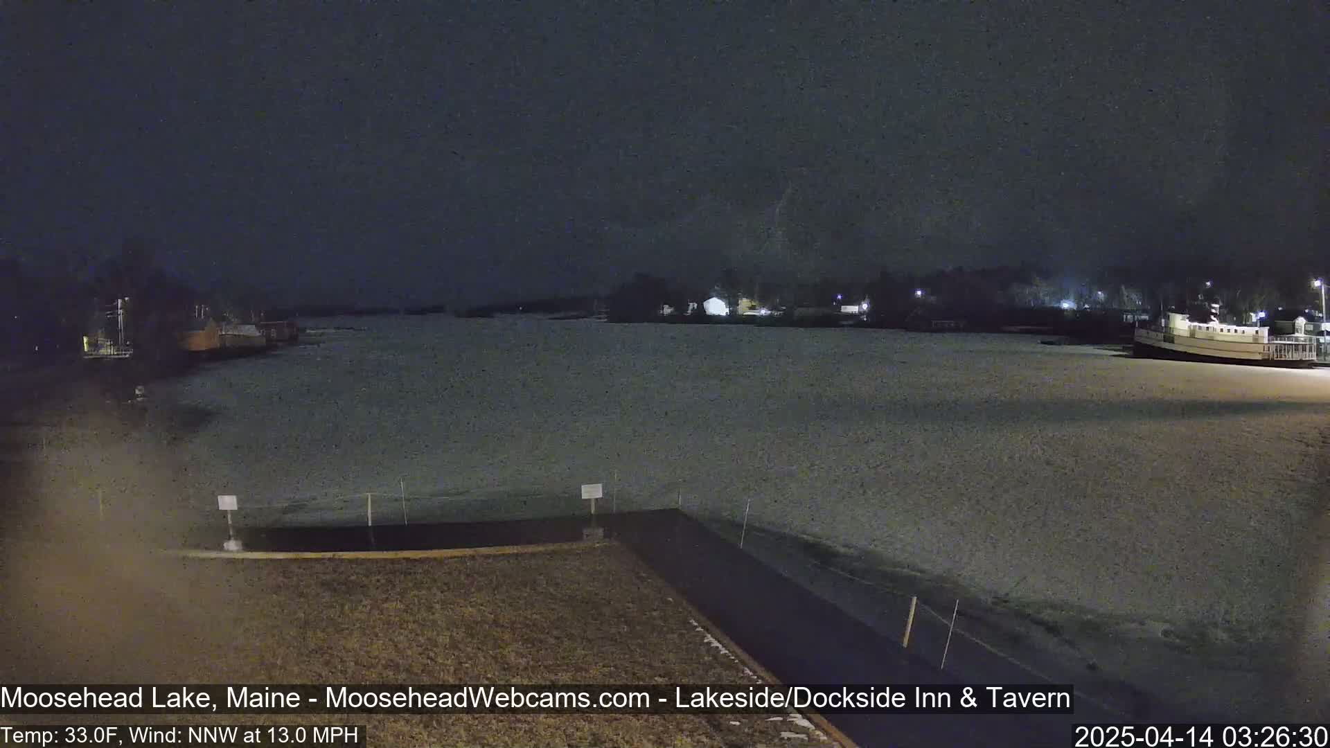A nighttime view of a snow-covered lake with buildings and a boat visible in the distance under a dark sky.