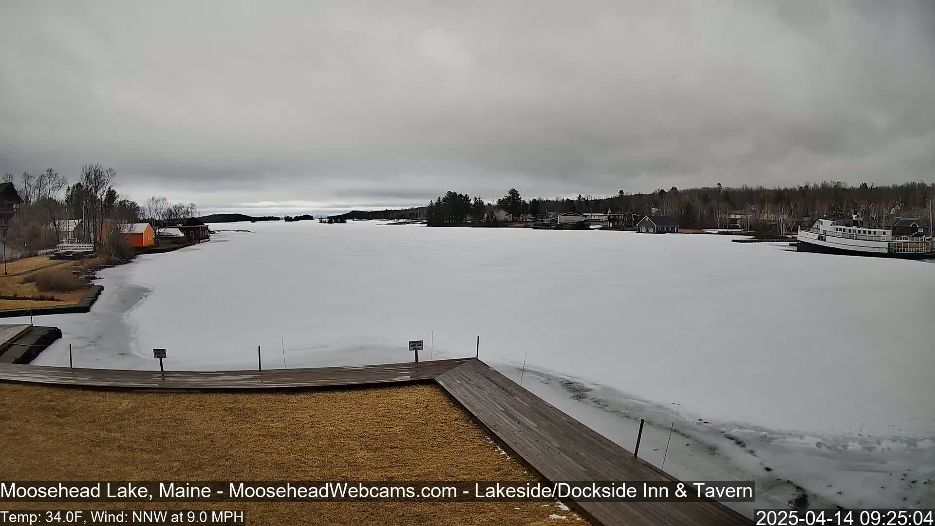 A mostly frozen lake is seen from a wooden dock, with a few buildings and a boat visible in the distance under an overcast sky.
