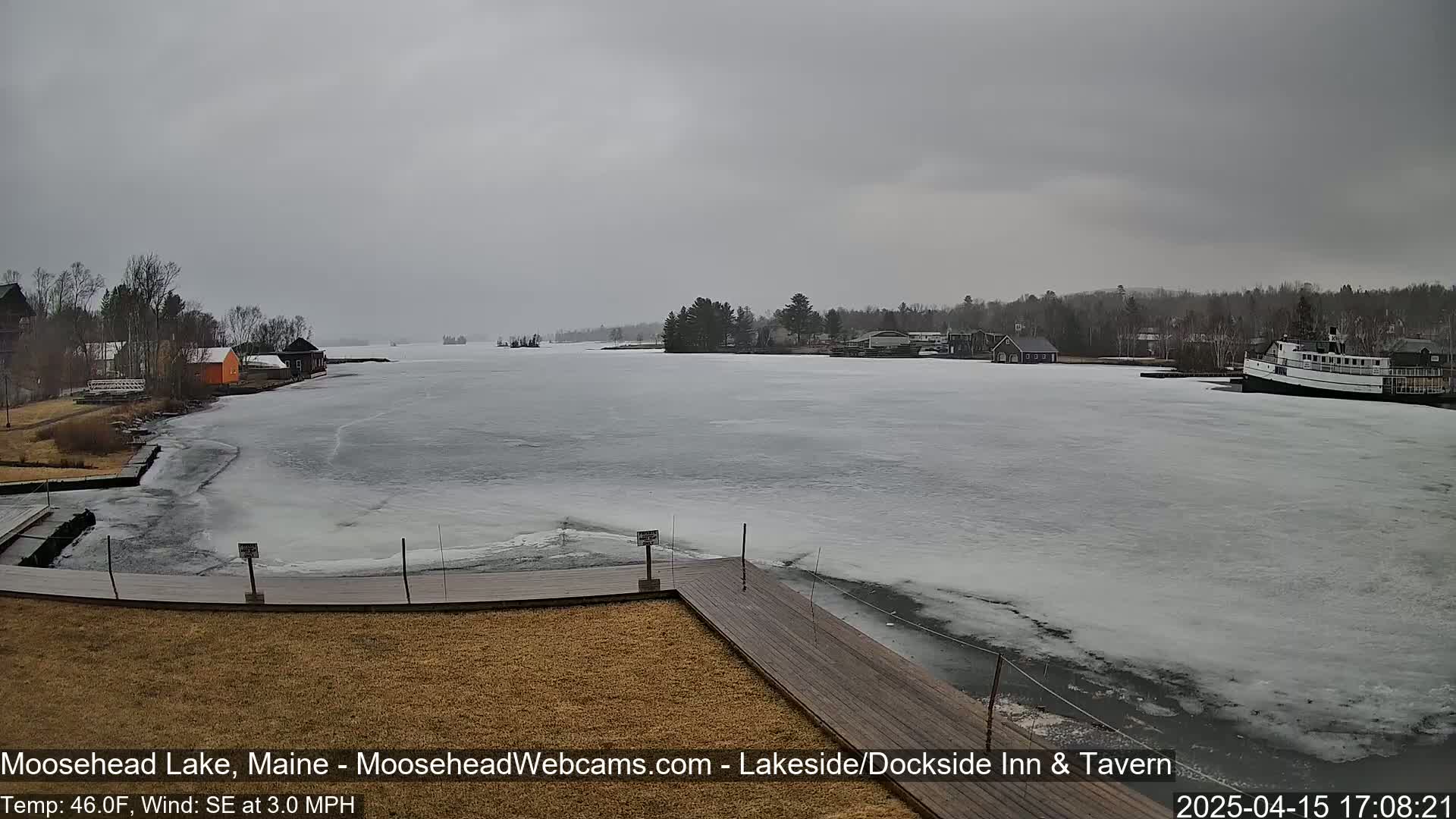 A partly frozen lake is viewed from a wooden dock, under an overcast sky.