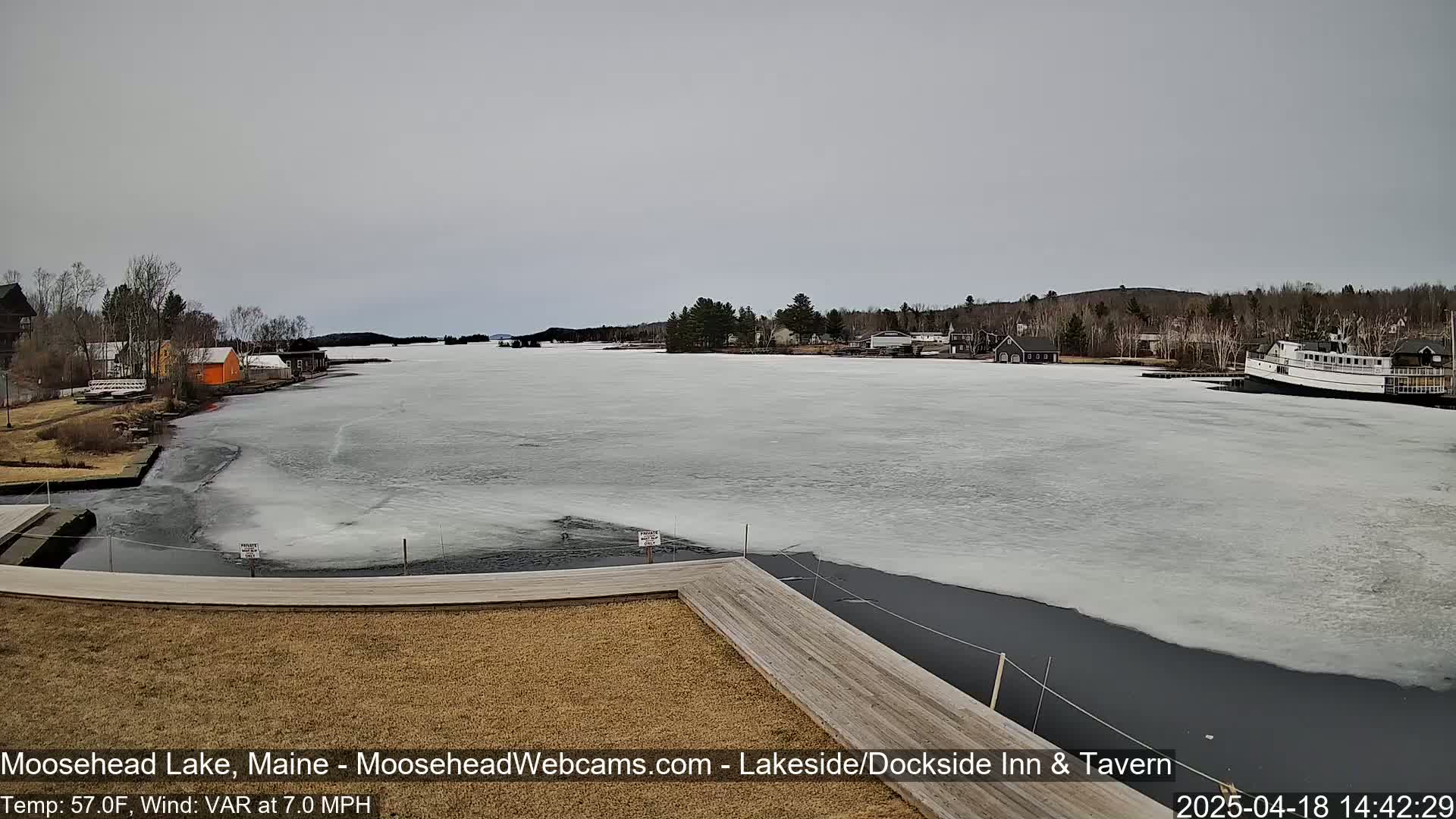 A partially frozen lake is seen from a wooden dock, with buildings and trees lining the shore under an overcast sky.