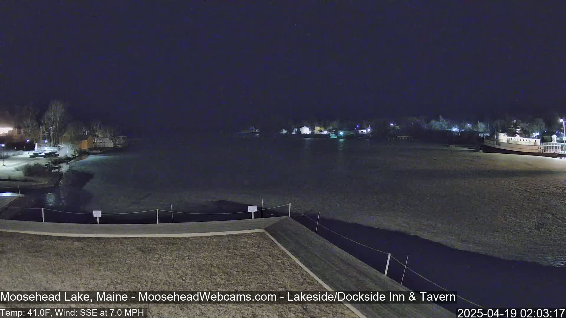 A nighttime view of a calm lake with a shoreline featuring buildings and a boat, under a dark sky with a SSE wind at 7.0 mph and a temperature of 41°F.