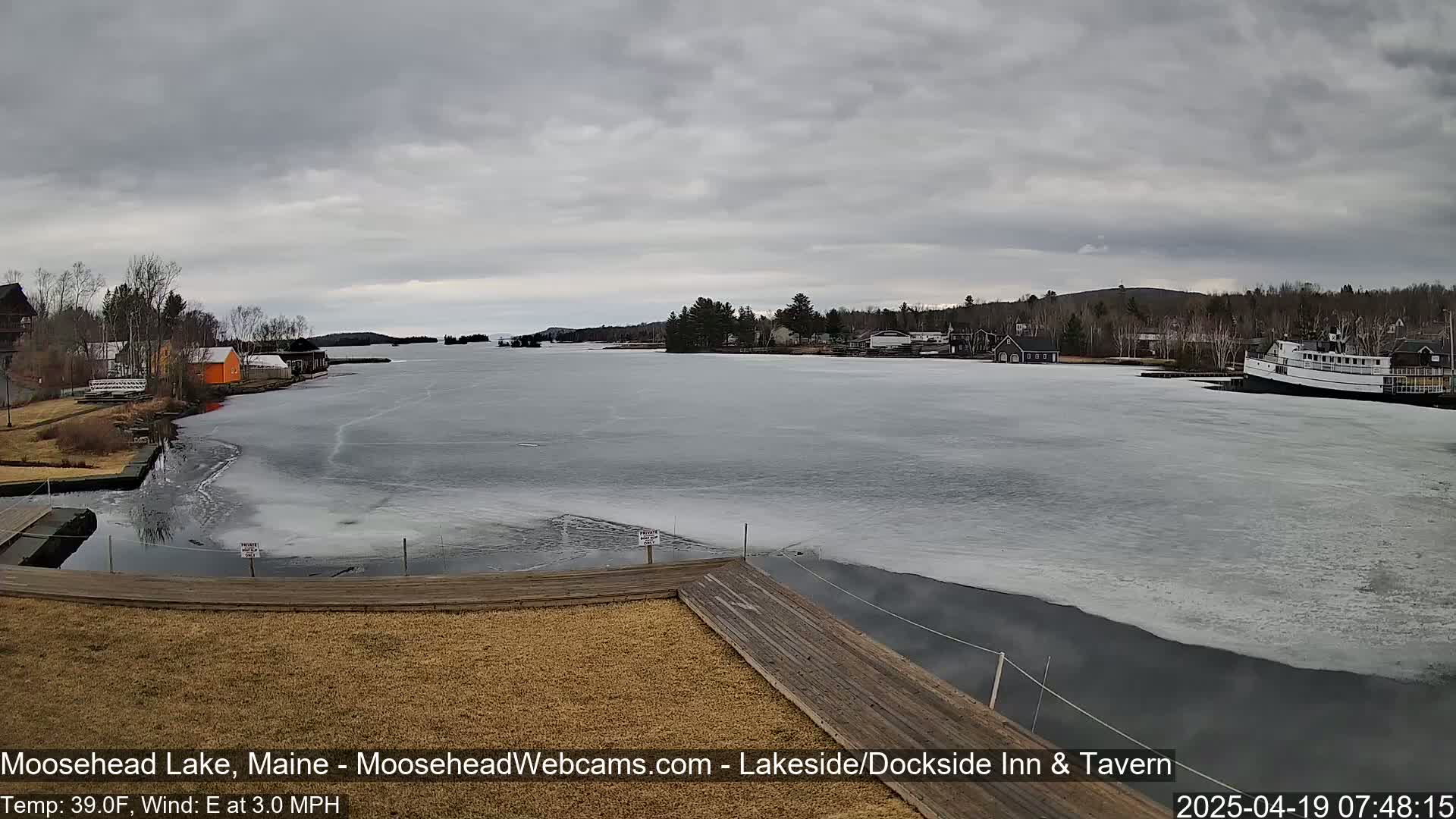 A partially frozen lake is seen from a dock, under an overcast sky, with buildings and a boat visible in the distance.