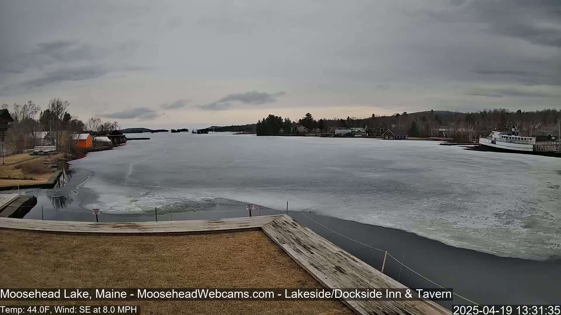 A partially frozen lake, under a gray sky, is seen from a wooden dock, with buildings and a large boat visible in the distance.